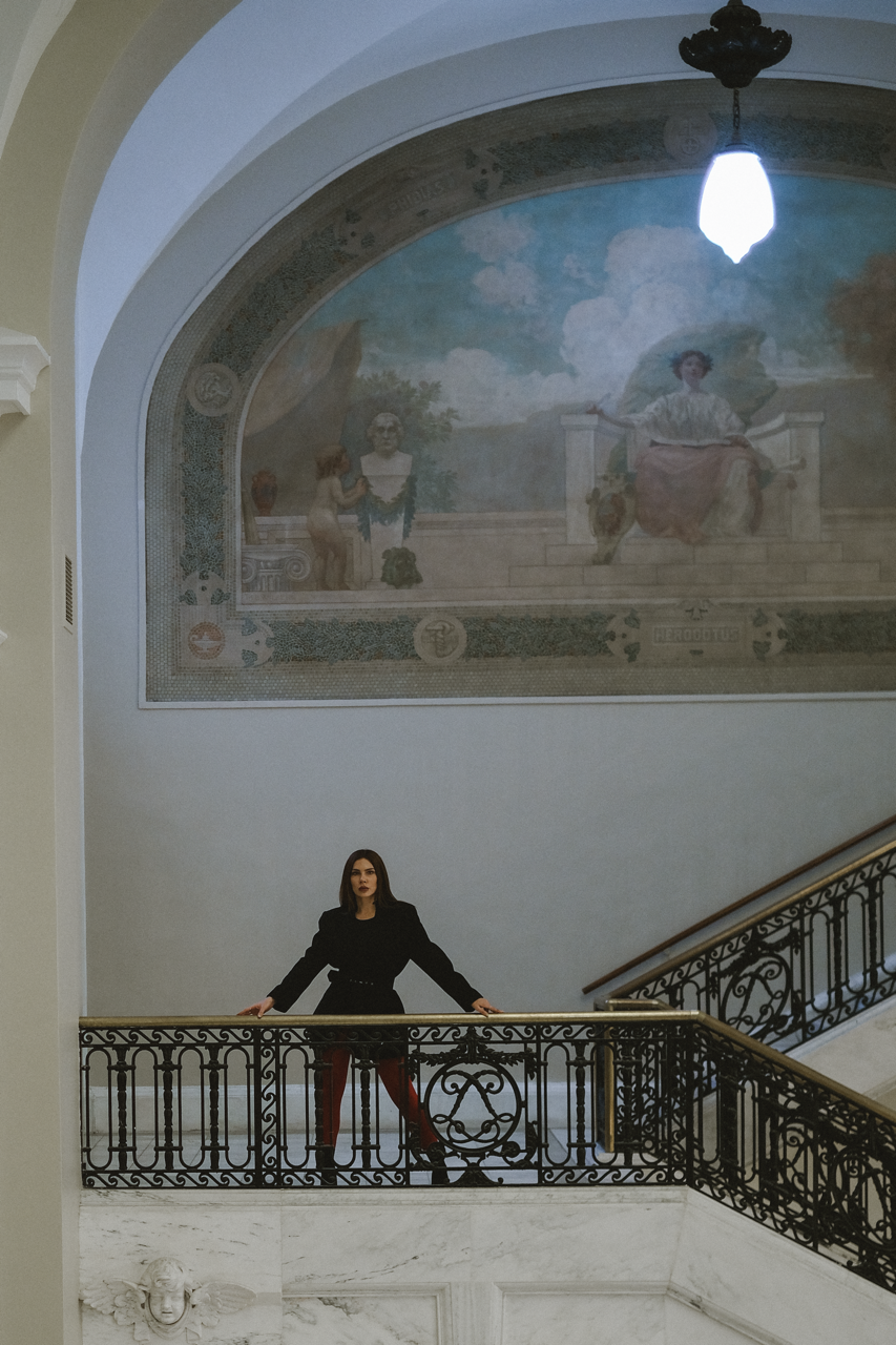 A woman with dark hair and dark lipstick standing on a curved stair railing in a grand building with a large mural on the wall behind her.