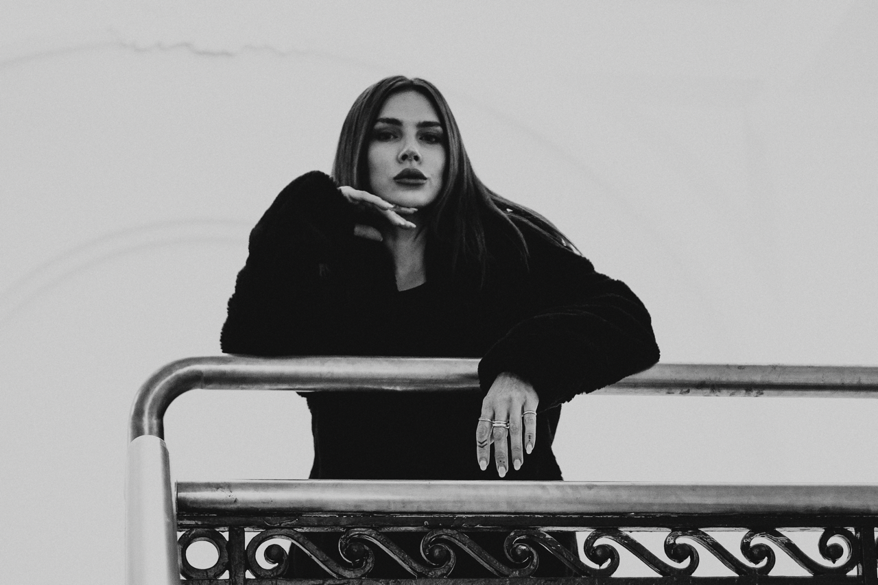 Black and white photo of a woman with long hair posing behind a metal railing, with her left arm resting on it and her right hand near her chin, wearing a dark coat.
