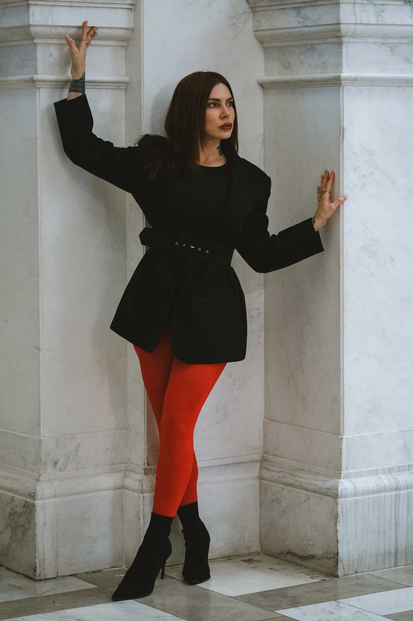 A woman with long dark hair and dark lipstick wearing a black blazer, red tights, and black high heels standing against a marble wall with her hands touching the wall and looking to the side.