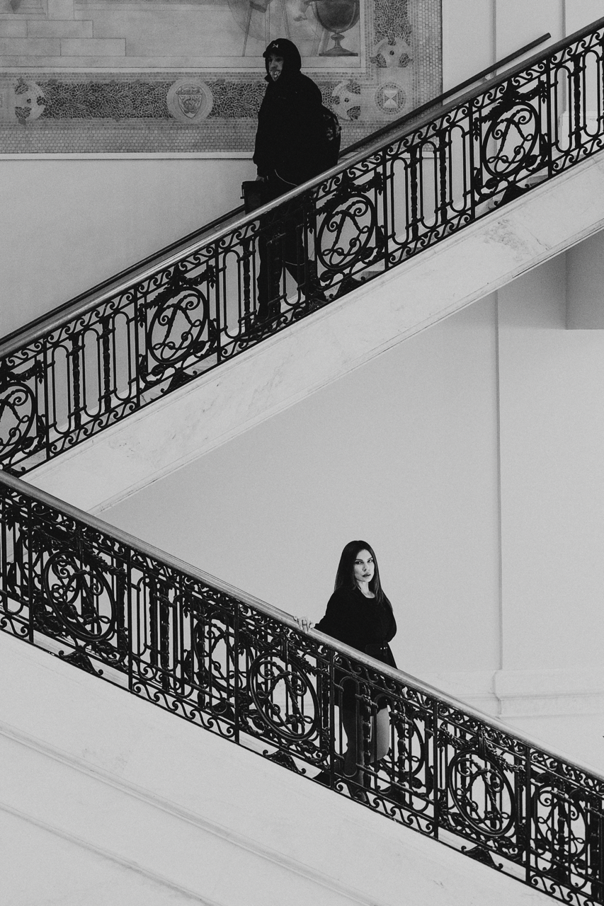 A black-and-white photo shows two women standing on different levels of a staircase with ornate black iron railings. The woman on the upper level is smiling and carrying a bag, while the woman on the lower level has a serious expression and holds a b