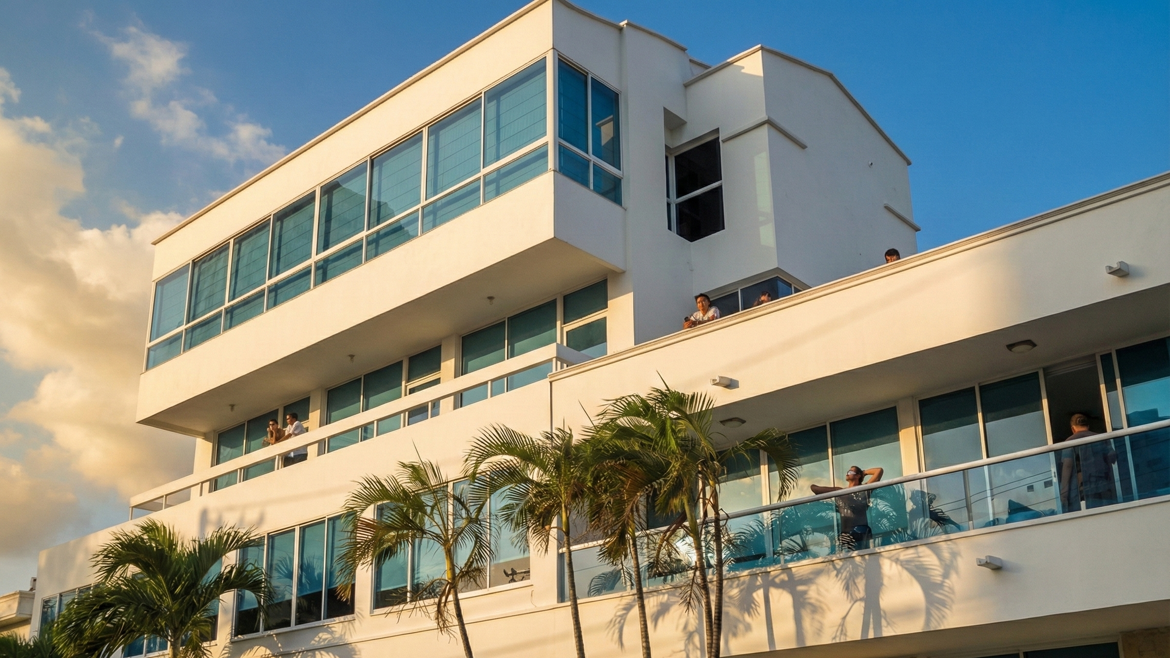 Edificio moderno de color blanco con varias terrazas y grandes ventanas, con palmeras en el primer plano y un cielo con algunas nubes al atardecer. Personas disfrutan en las terrazas.