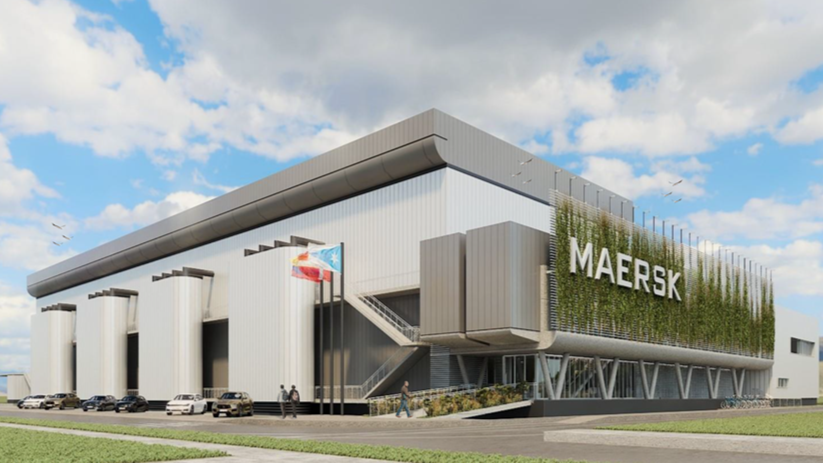 Vista frontal de un edificio modernista con el nombre 'MAERSK' en un lado, con banderas en el área de entrada, varias personas y autos estacionados en el frente, y un cielo parcialmente nublado.
