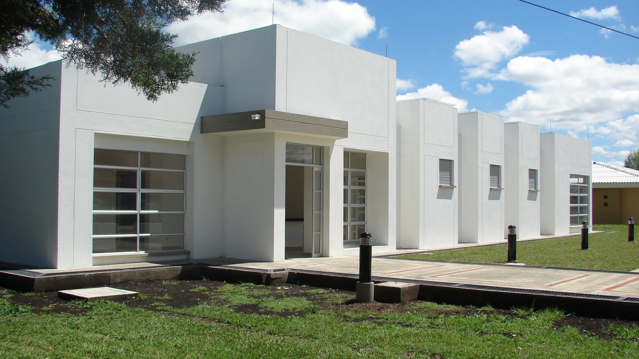 Edificio moderno de color blanco con ventanas grandes y barredora en la entrada, rodeado de césped y bajo cielo parcialmente nublado.