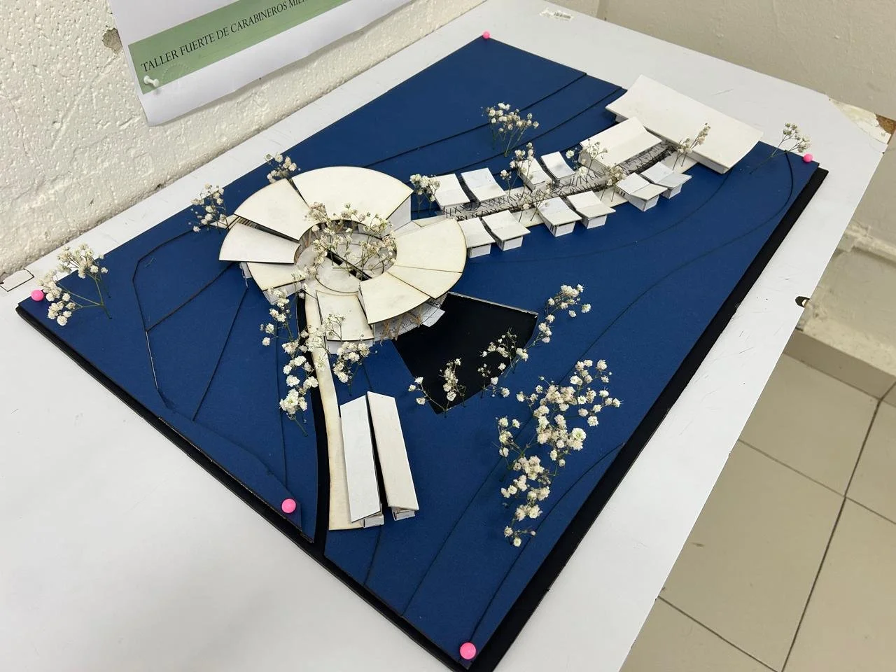 Maqueta arquitectónica de un edificio en un terreno con césped, con detalles en blanco y azules, decorada con pequeñas flores blancas.