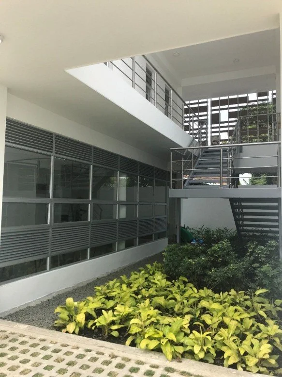 Vista interna de la Estación de Policía Cero Energía en Florida, Valle, Colombia. Un edificio moderno con ventanas de vidrio y una escalera metálica exterior, con áreas verdes y plantas en primer plano.