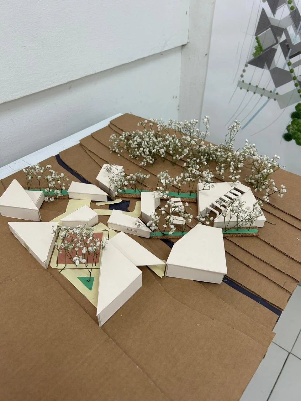 Maqueta arquitectónica de un complejo con edificios blancos y áreas verdes con árboles pequeños, colocada sobre una superficie de cartón.