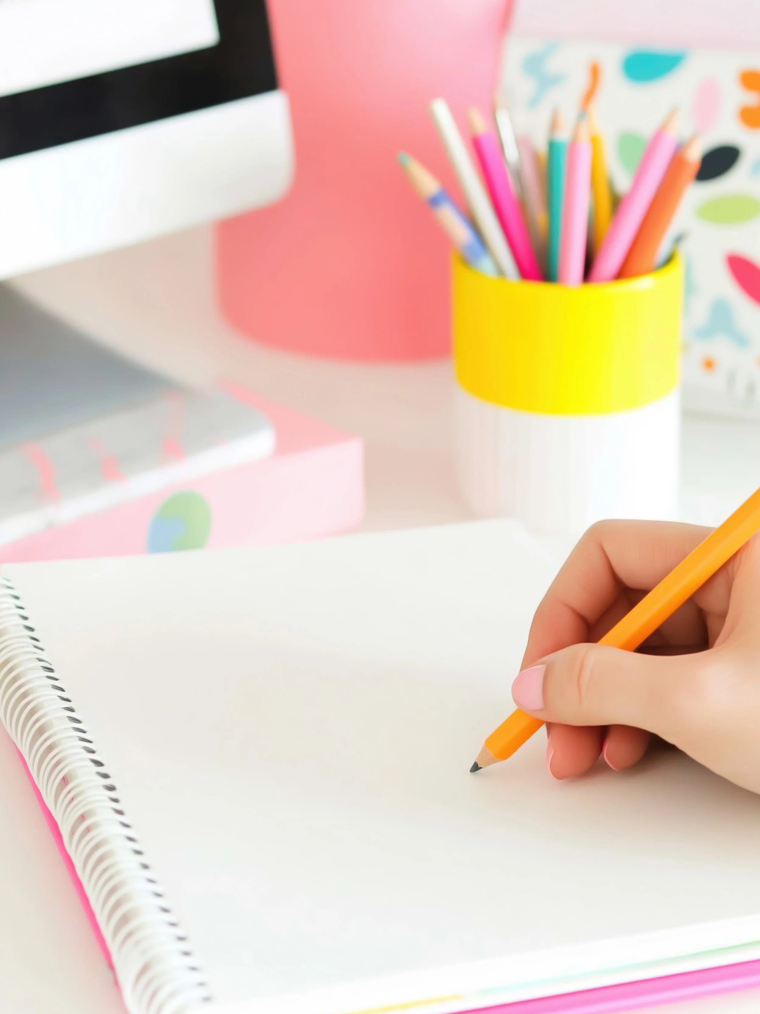 A person is holding an orange pencil and writing on an open white notebook with a black spiral binding. In the background, there are colorful pencils in a yellow and white cup, along with a pink container and a decorative box with colorful patterns.
