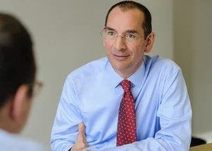 A man in a light blue dress shirt and red tie smiling during a conversation with another person.