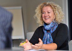 A woman with curly blonde hair smiling and talking to someone at a desk in an office.