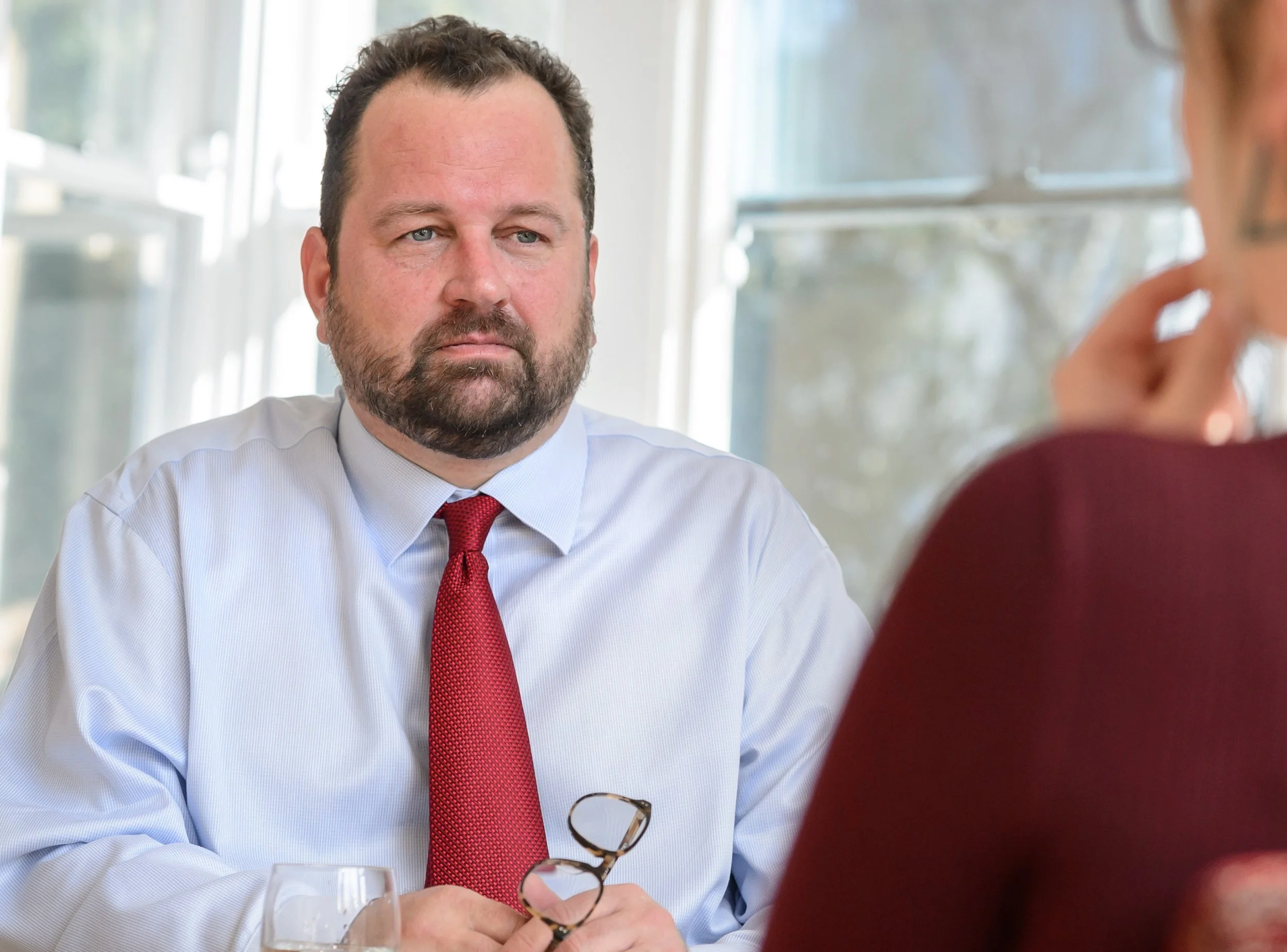 A serious-looking man with a beard, wearing a white shirt and red tie, holding reading glasses, seated at a table during an interview or meeting.