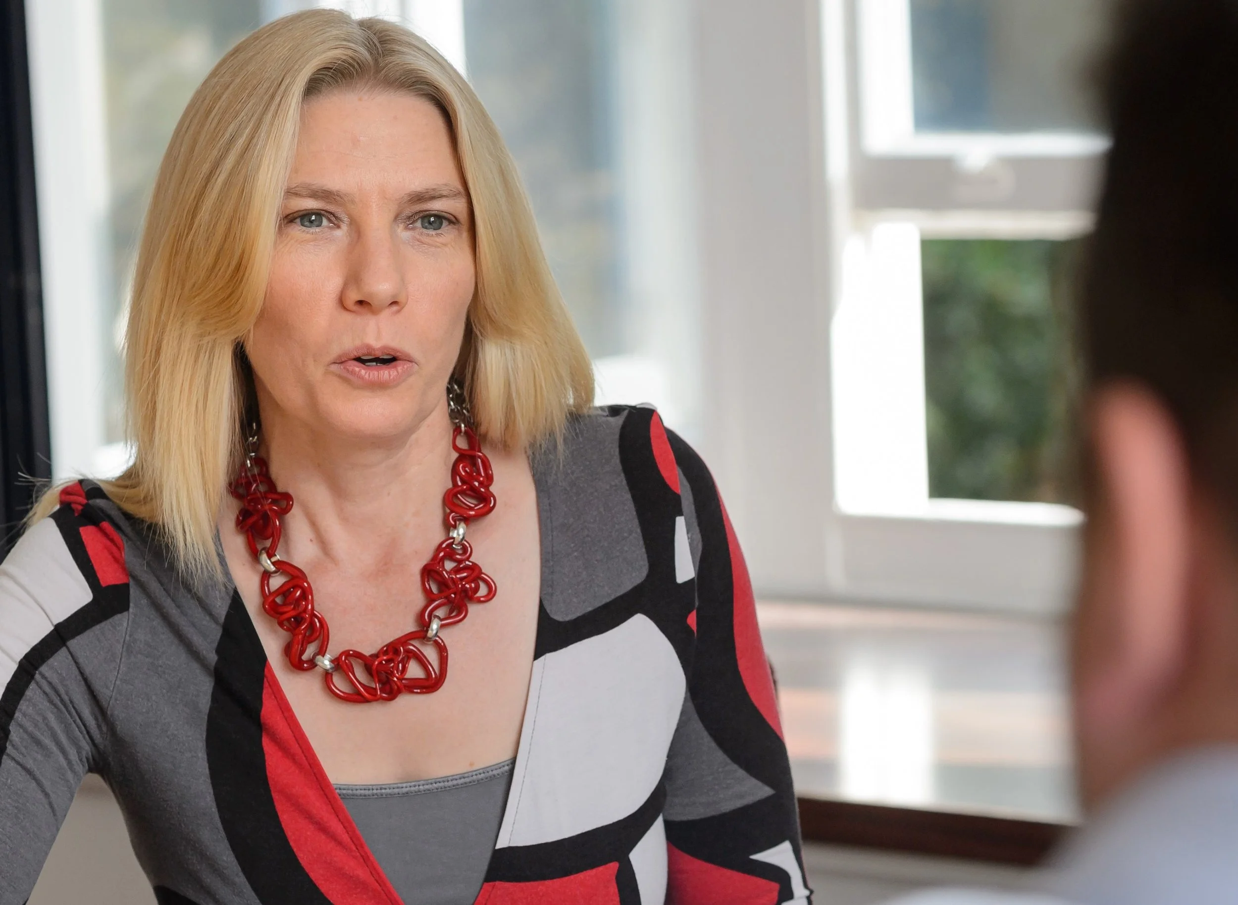 A blonde woman wearing a red and black patterned dress and a red statement necklace talking to a person whose back is visible in the foreground. They are in a room with large windows.