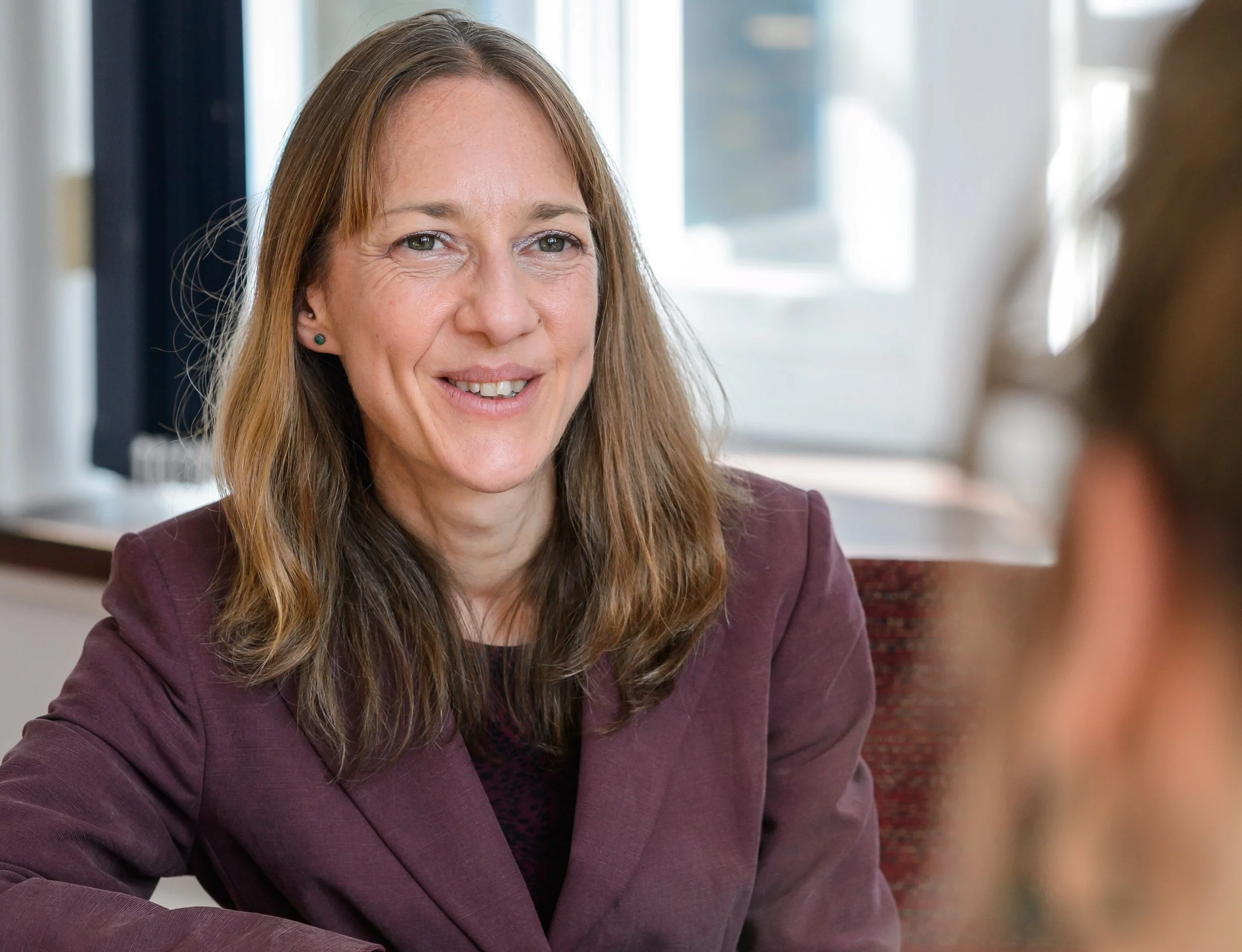 A woman with brown hair, wearing a maroon blazer, smiling and talking to another person in an indoor setting.