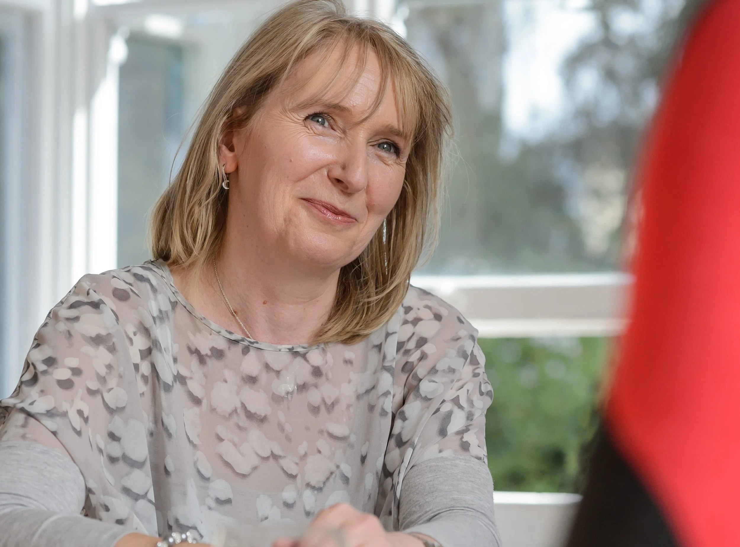A woman with shoulder-length blonde hair and blue eyes smiling while looking at someone off-camera, sitting near a window with a blurred background.