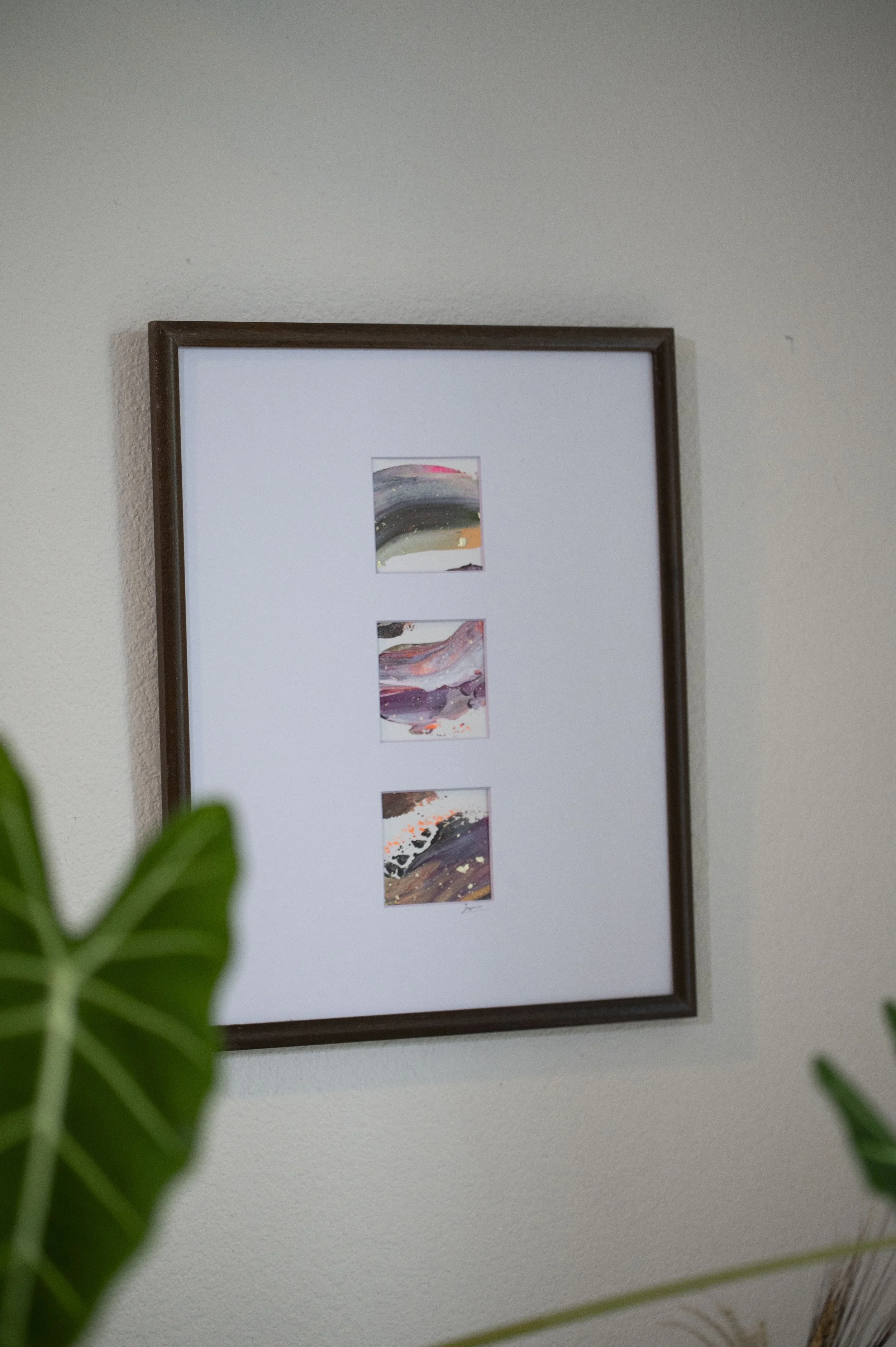 Framed artwork with three abstract paintings on white background, partially obscured by green leaves in the foreground.