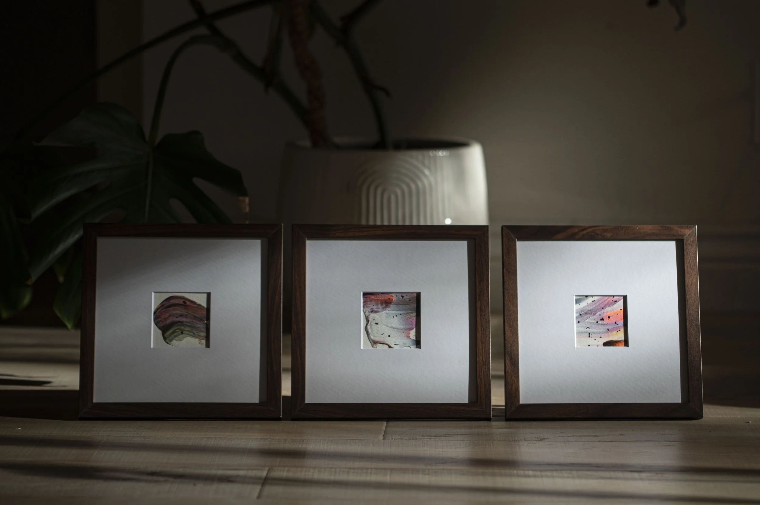Three framed abstract artworks in small square sizes, displayed on a wooden surface, with a dark background and a white vase with a plant behind them.
