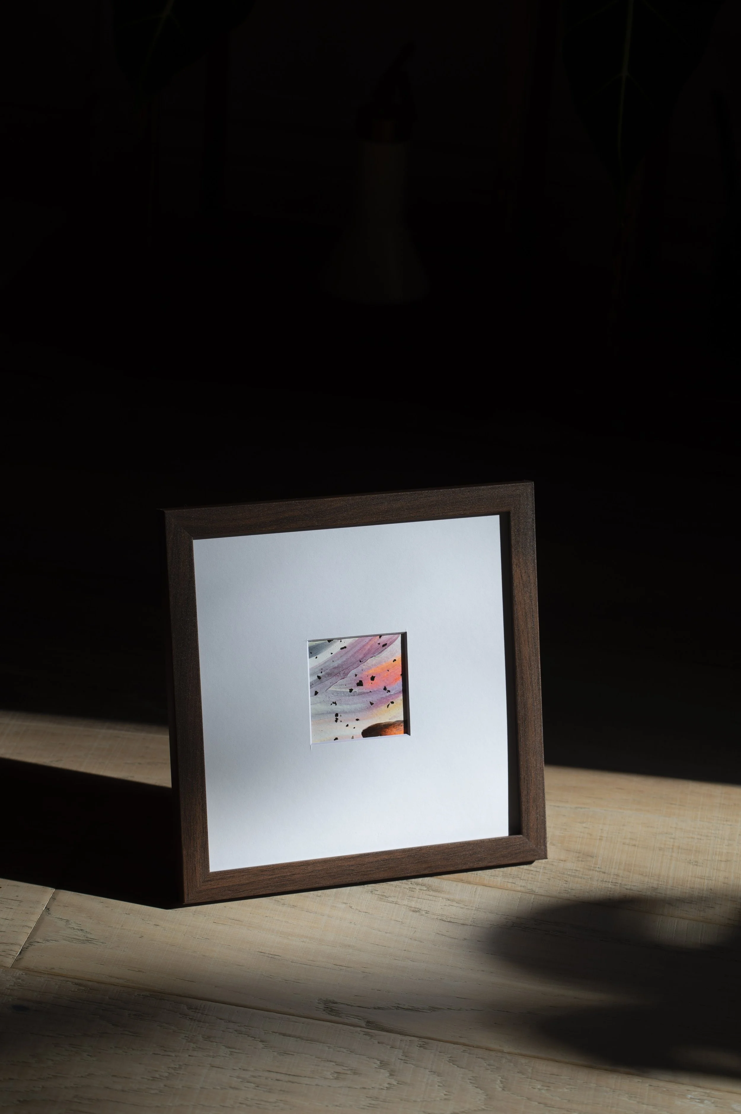 A small framed picture with a colorful sunset or sunrise scene, placed on a wooden surface with a dark background and a spotlight casting a shadow.