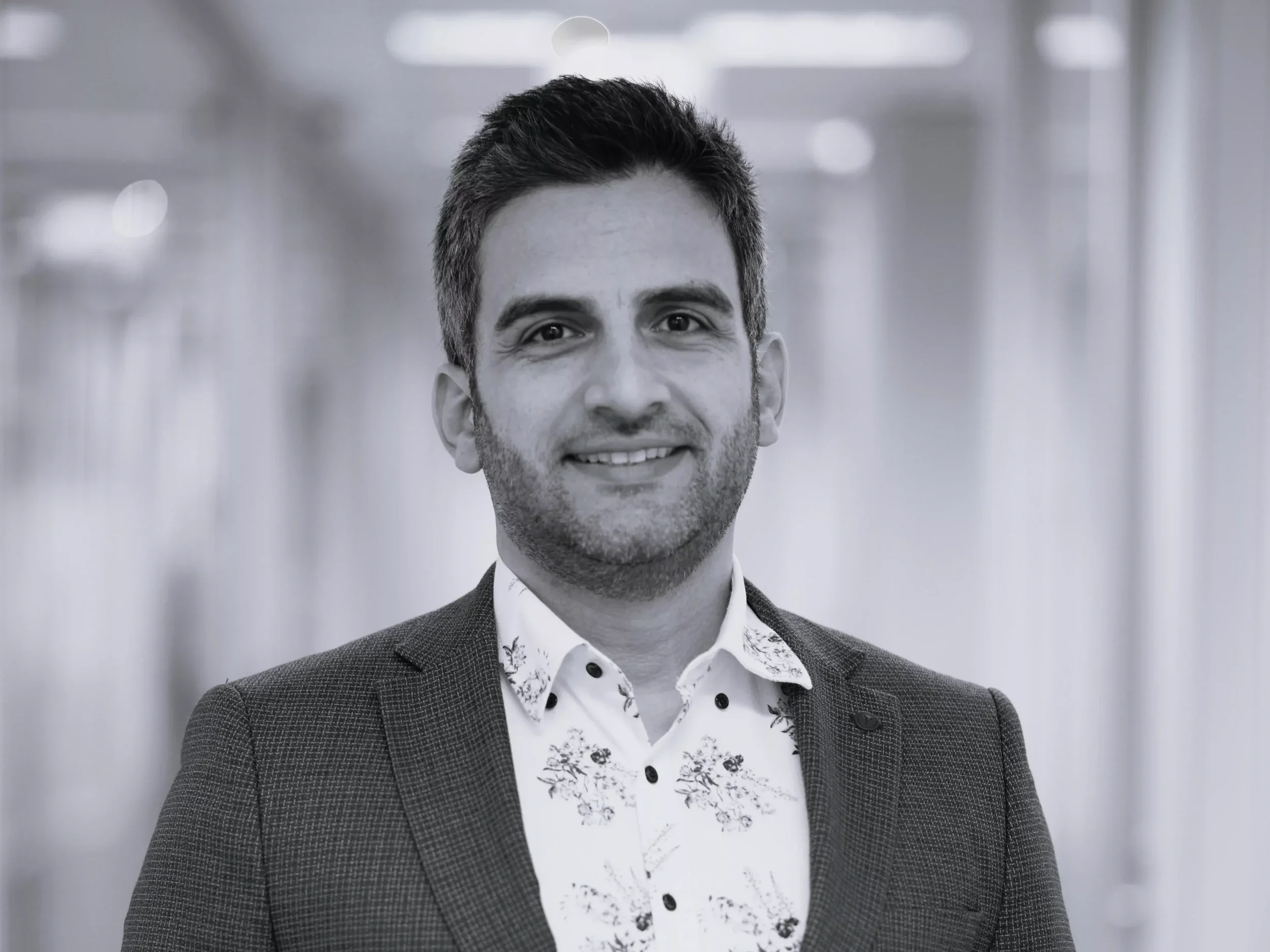 A professional man with dark hair and a beard, wearing a floral shirt and a blazer, smiling at the camera in an office corridor.