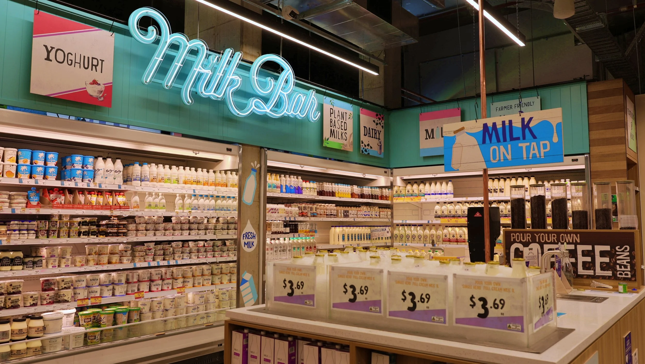 A dairy section in a grocery store with a glowing neon sign that says 'Milk Bar', shelves filled with yogurt containers, and a tap station for pouring fresh milk. Signage indicates plant-based milks and milk on tap, with a tip jar for coffee beans.
