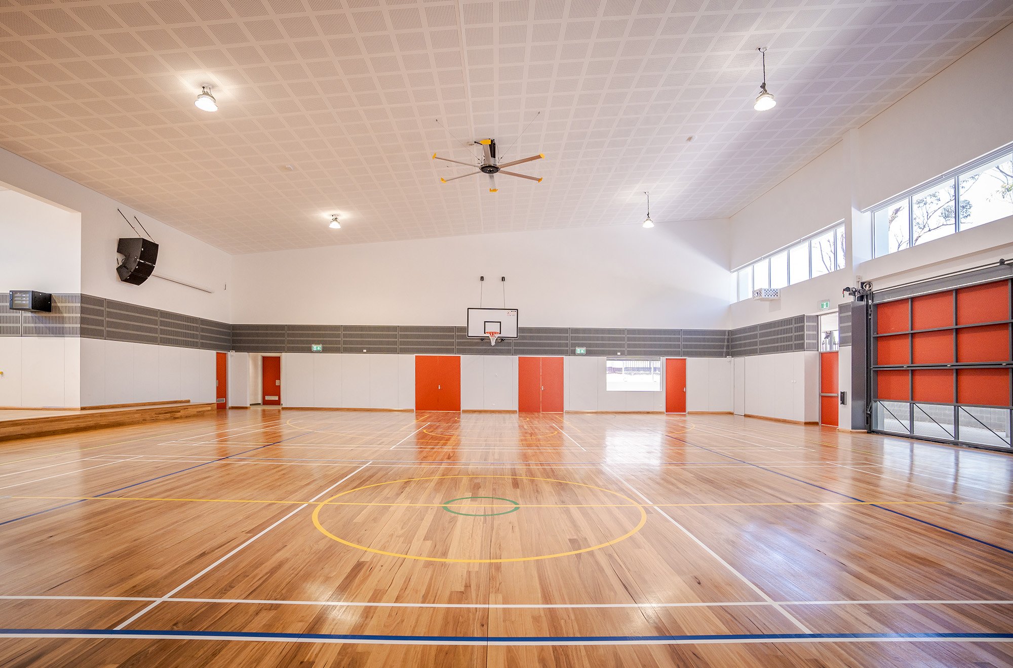 An empty indoor basketball court with a wooden floor, a basketball hoop, and a large garage-style door.