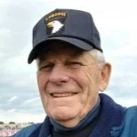 Older man wearing a Navy cap and a dark blue jacket, smiling outdoors with a cloudy sky in the background.