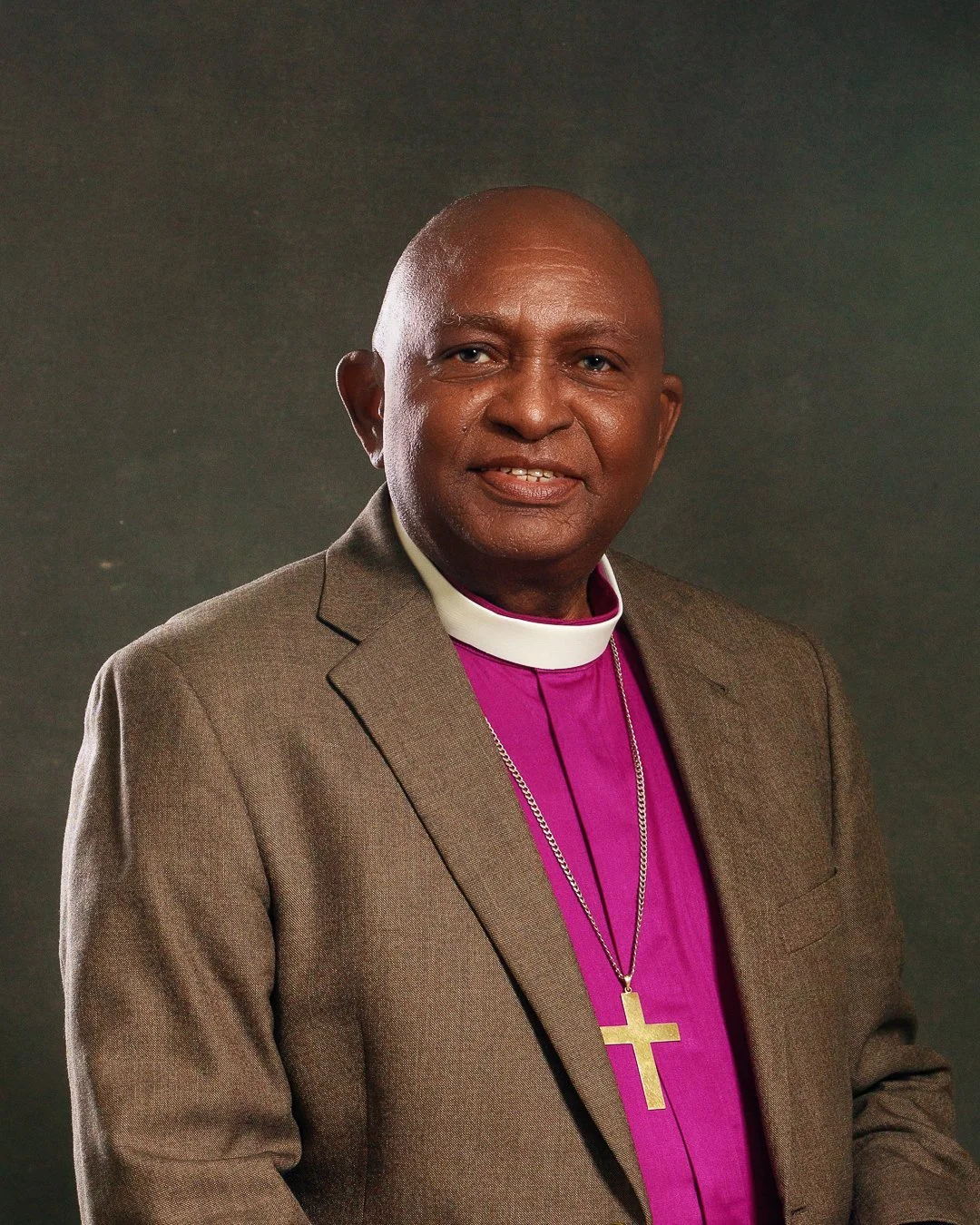 A man dressed in a suit and clergy attire, wearing a purple clerical shirt with a white collar and a cross necklace, smiling at the camera against a plain dark background.