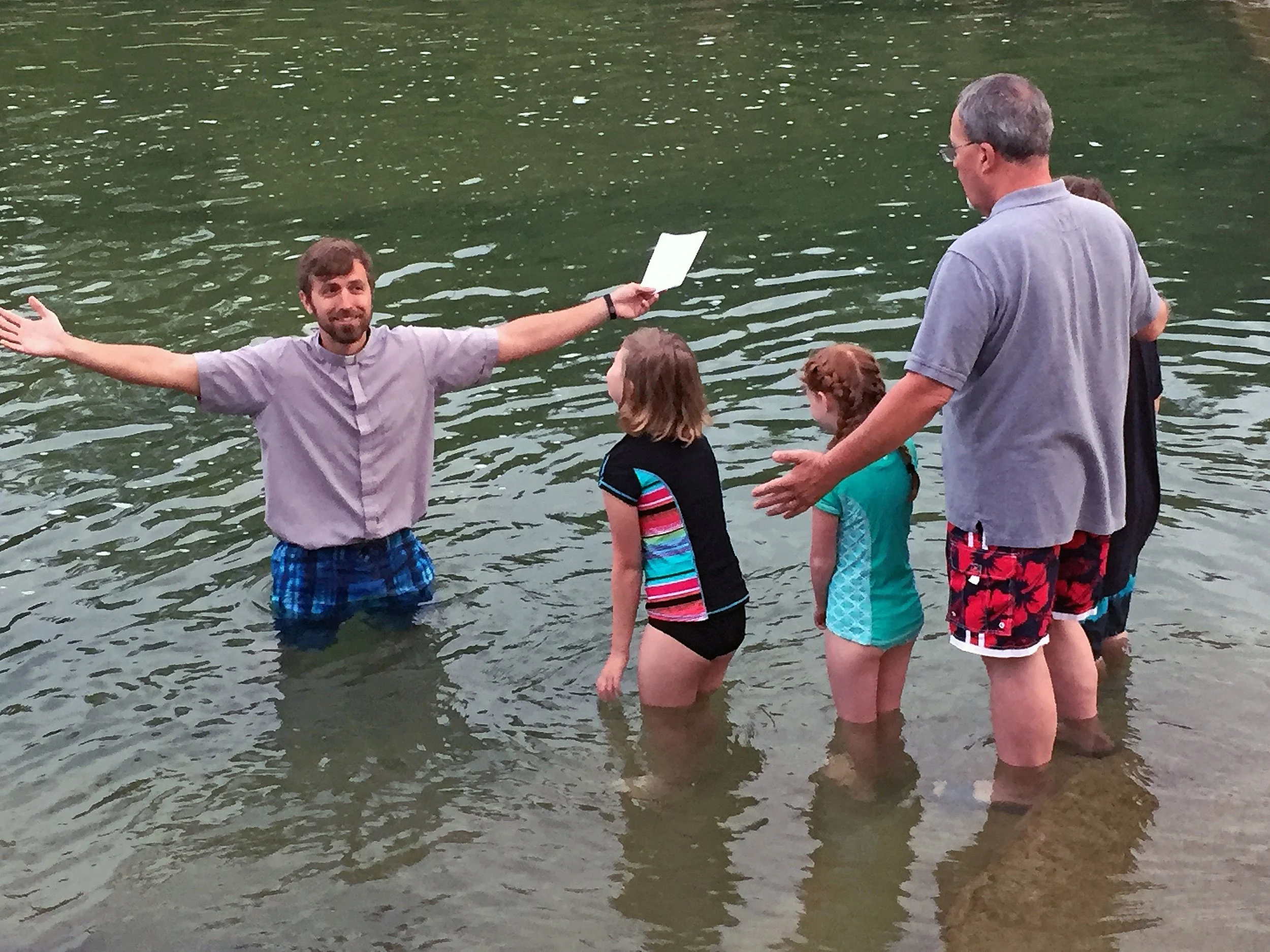 A baptism ceremony taking place in a lake with a man in a purple shirt and blue shorts baptizing three children, while another man in a gray shirt and red shorts stands nearby. One young girl is in black swimwear with colorful stripes, and two girls are in blue swimsuits.