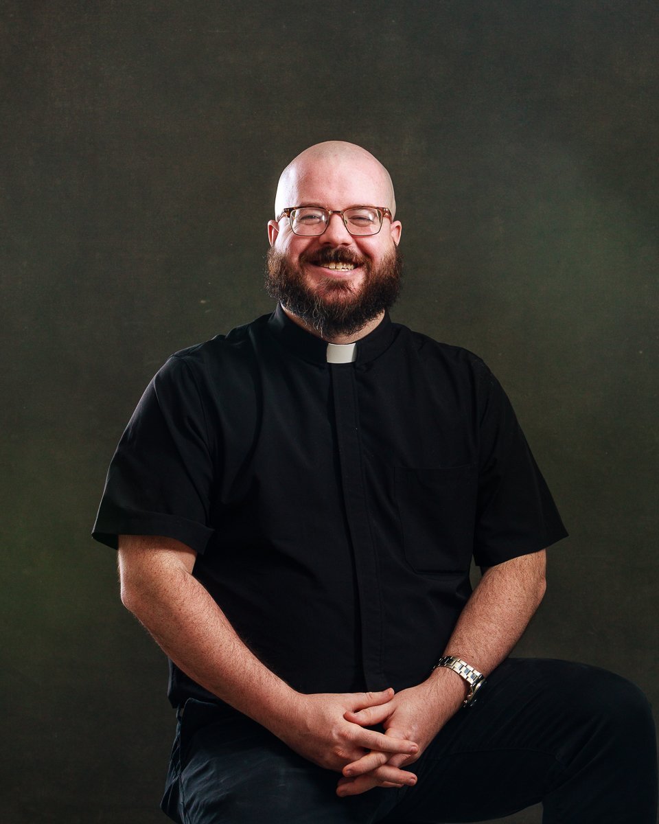 A smiling man with glasses and a beard, dressed in a black clerical shirt with a white collar, sitting against a dark background.