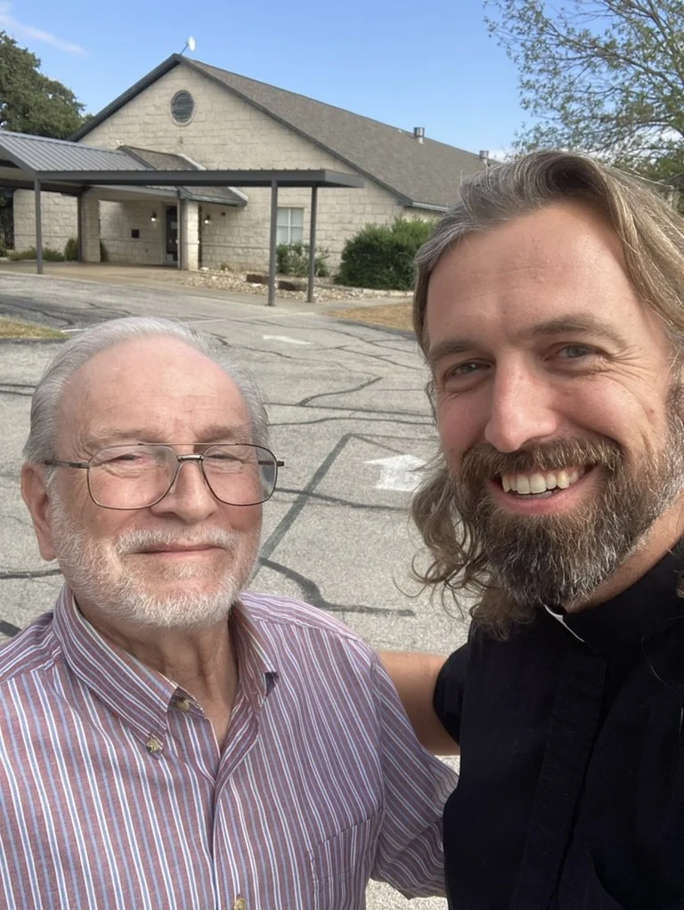 Two men smiling outside in front of a building with a parking lot. One is an elderly man with glasses and a striped shirt, the other is a younger man with long hair and a beard, wearing a black shirt.