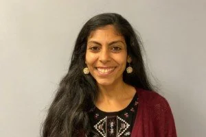 A woman with long dark hair wearing earrings and a maroon cardigan smiles at the camera against a plain light-colored background.