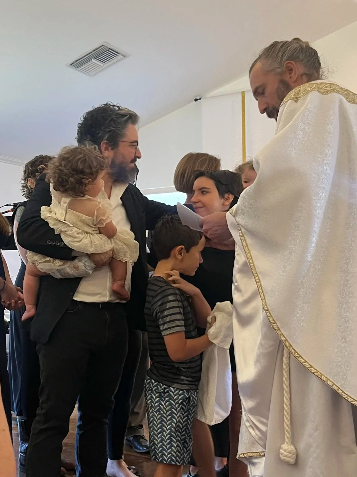 A religious ceremony, likely a baptism or first communion, taking place indoors with children and adults gathered around a priest dressed in white robes. A young boy is participating in the ceremony, with others watching. The setting appears to be a church or a religious venue.