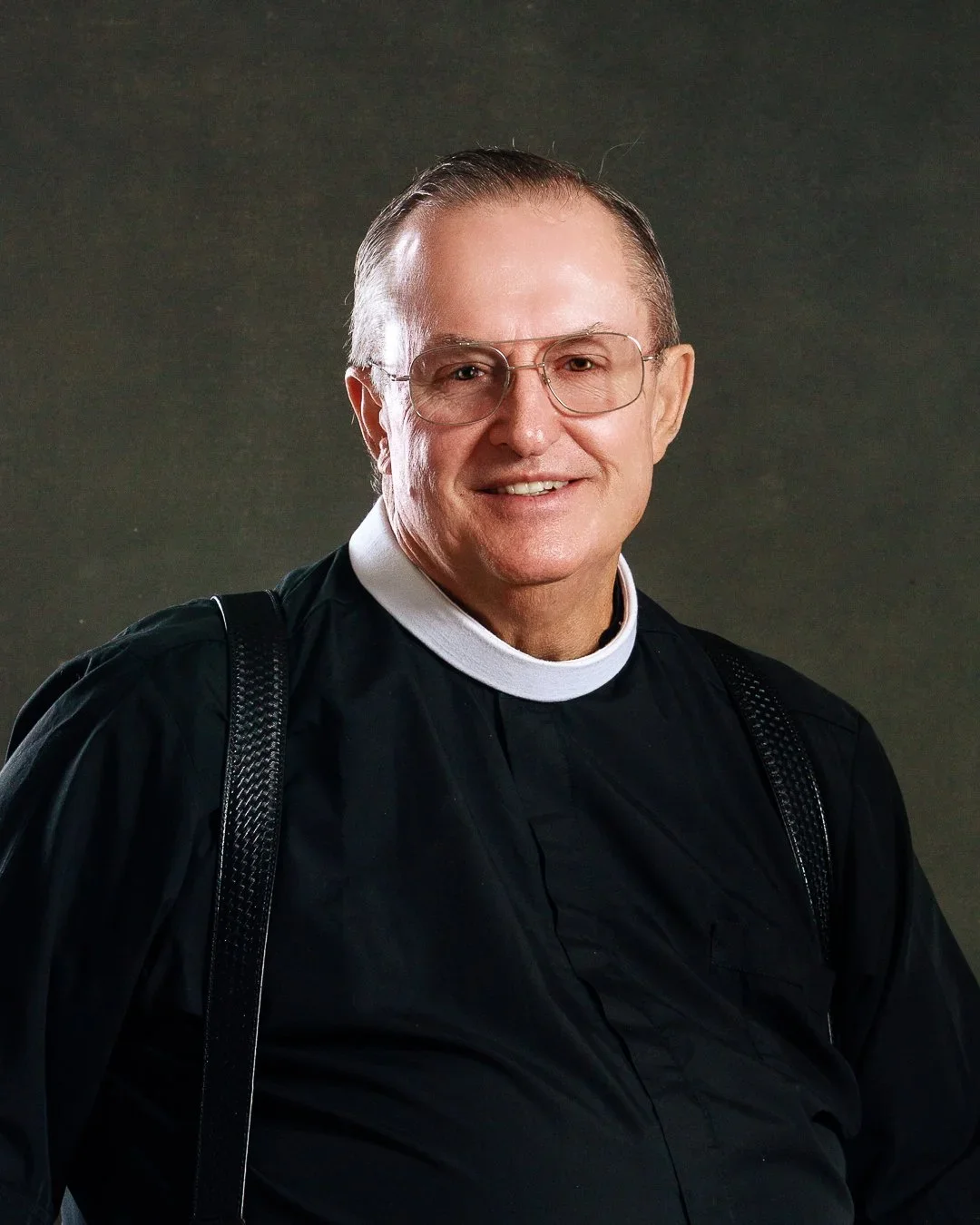 Portrait of a middle-aged man with glasses, wearing a black clerical shirt with a white collar, smiling against a dark background.