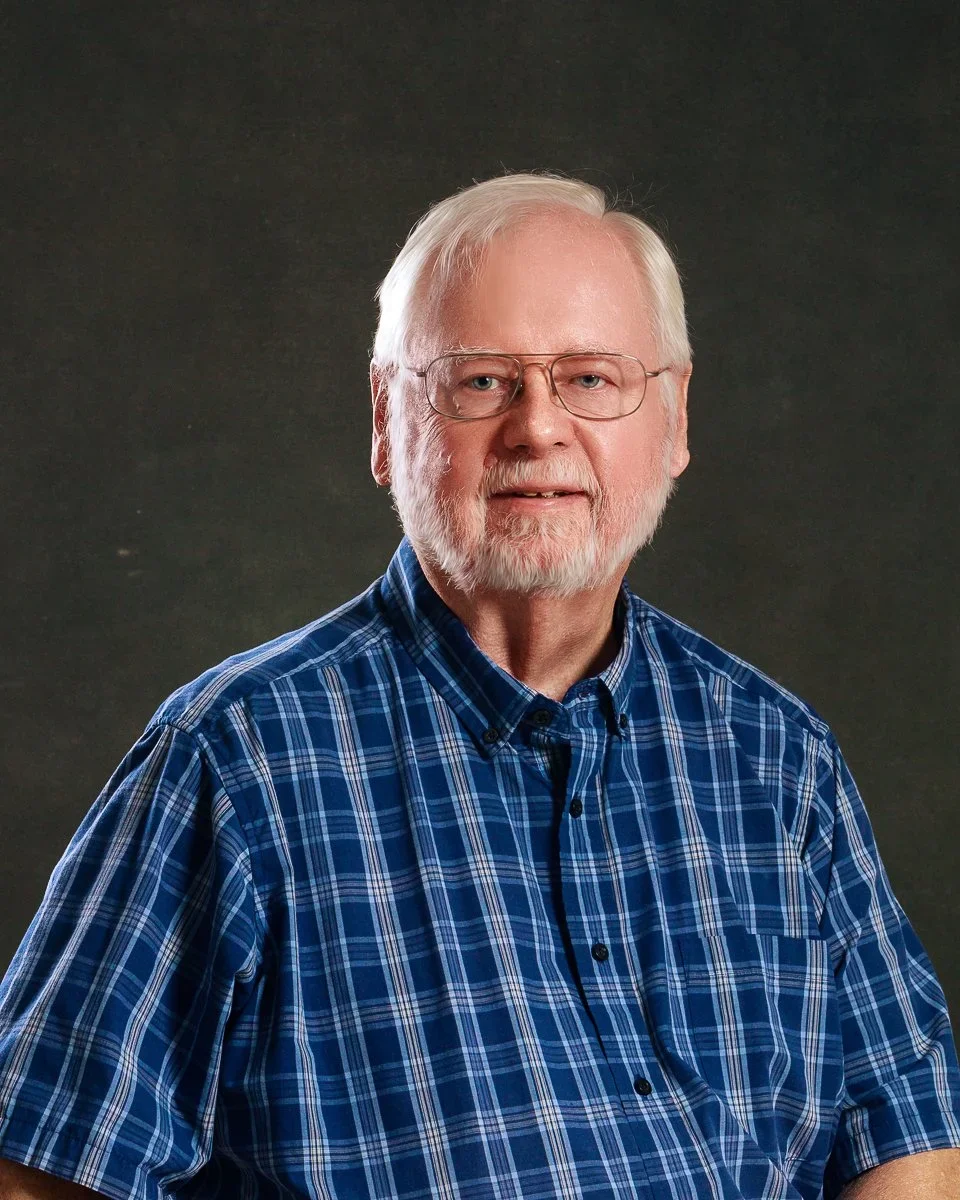 Portrait of an older man with white hair, beard, and glasses, wearing a blue plaid shirt, against a dark background.