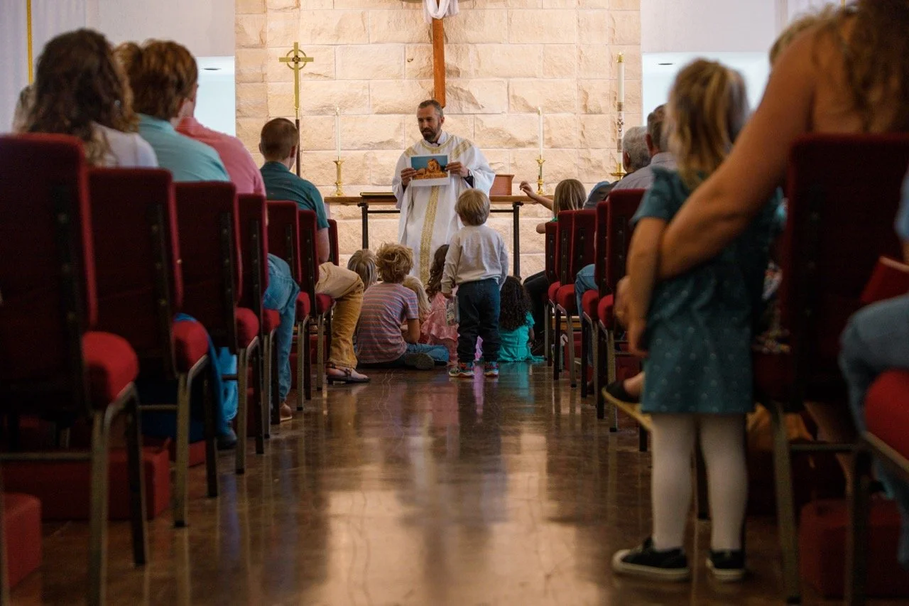 A priest leading a church service in a sanctuary with children seated on the floor and adults in red chairs on either side.