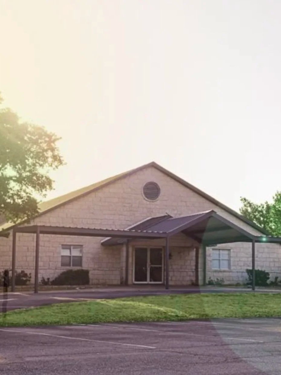 A single-story beige brick building with a pitched roof and a small circular window near the roof peak. There is a covered entryway with a metal frame and a glass door. The building is surrounded by a parking lot and some green grass and trees.