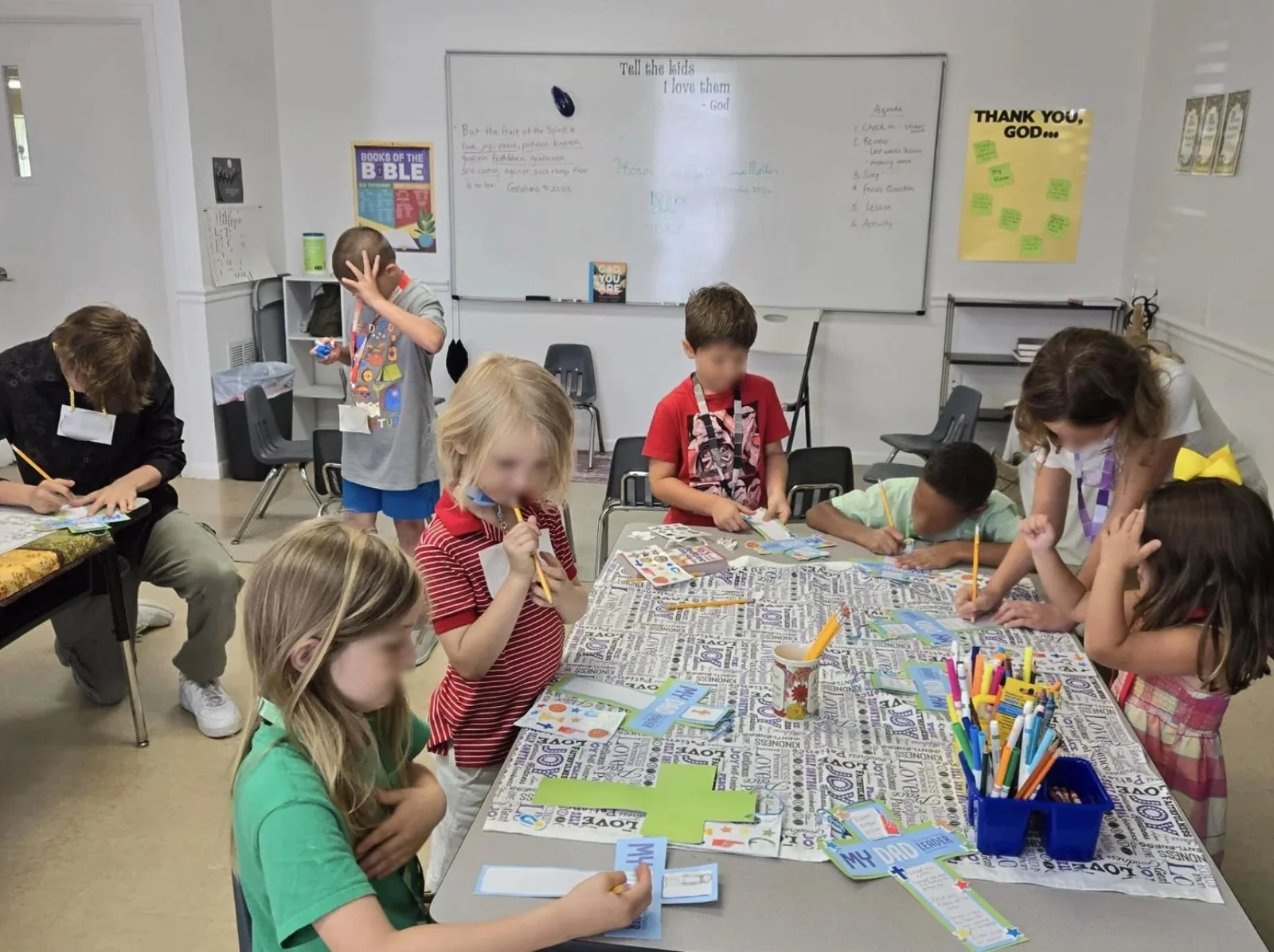 Children and a teacher gathered around a table in a classroom, engaging in a craft or learning activity with colorful papers, markers, and cards.