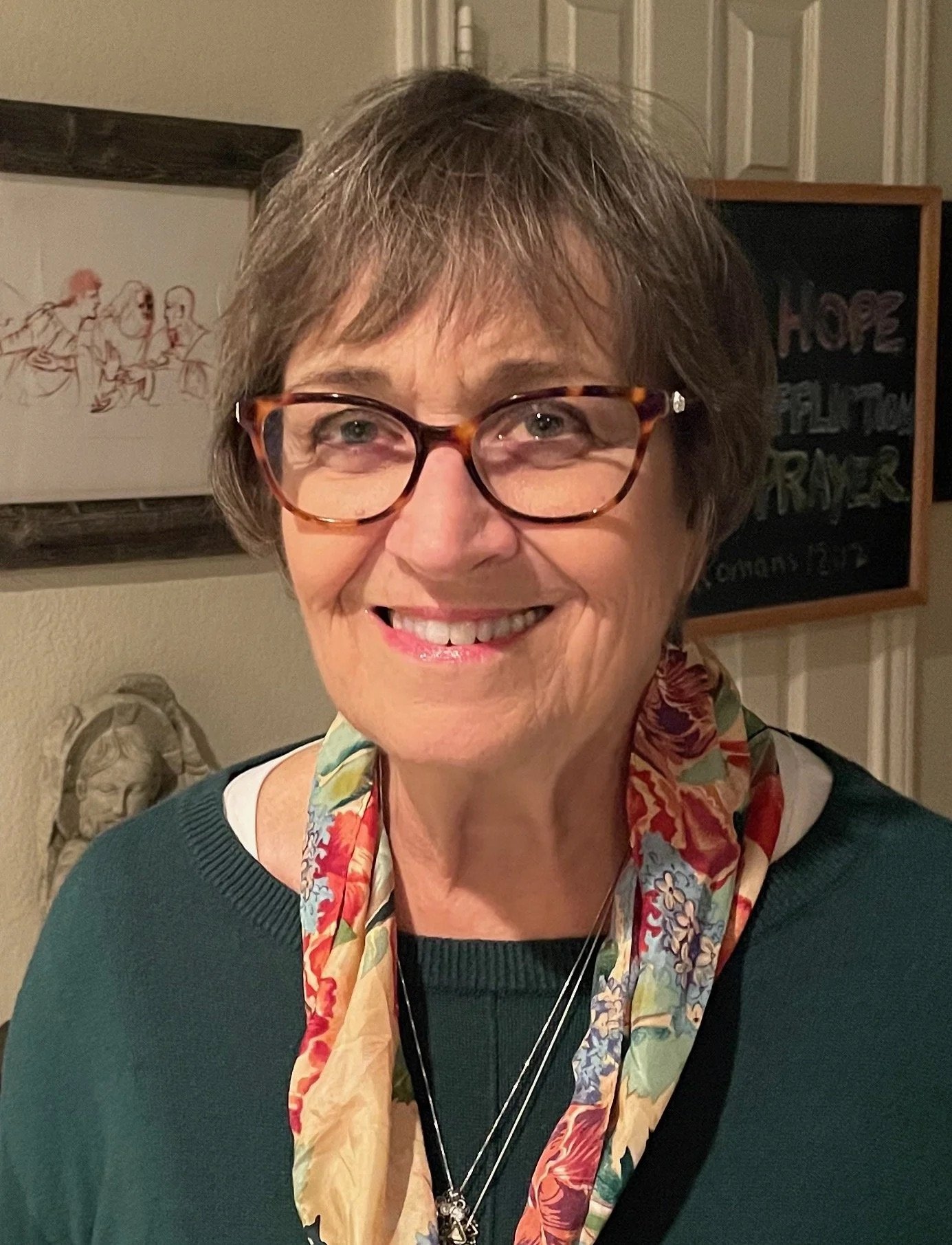 A smiling older woman with short brown hair, wearing glasses, a dark green sweater, and a colorful floral scarf, standing indoors with framed artwork and a chalkboard in the background.