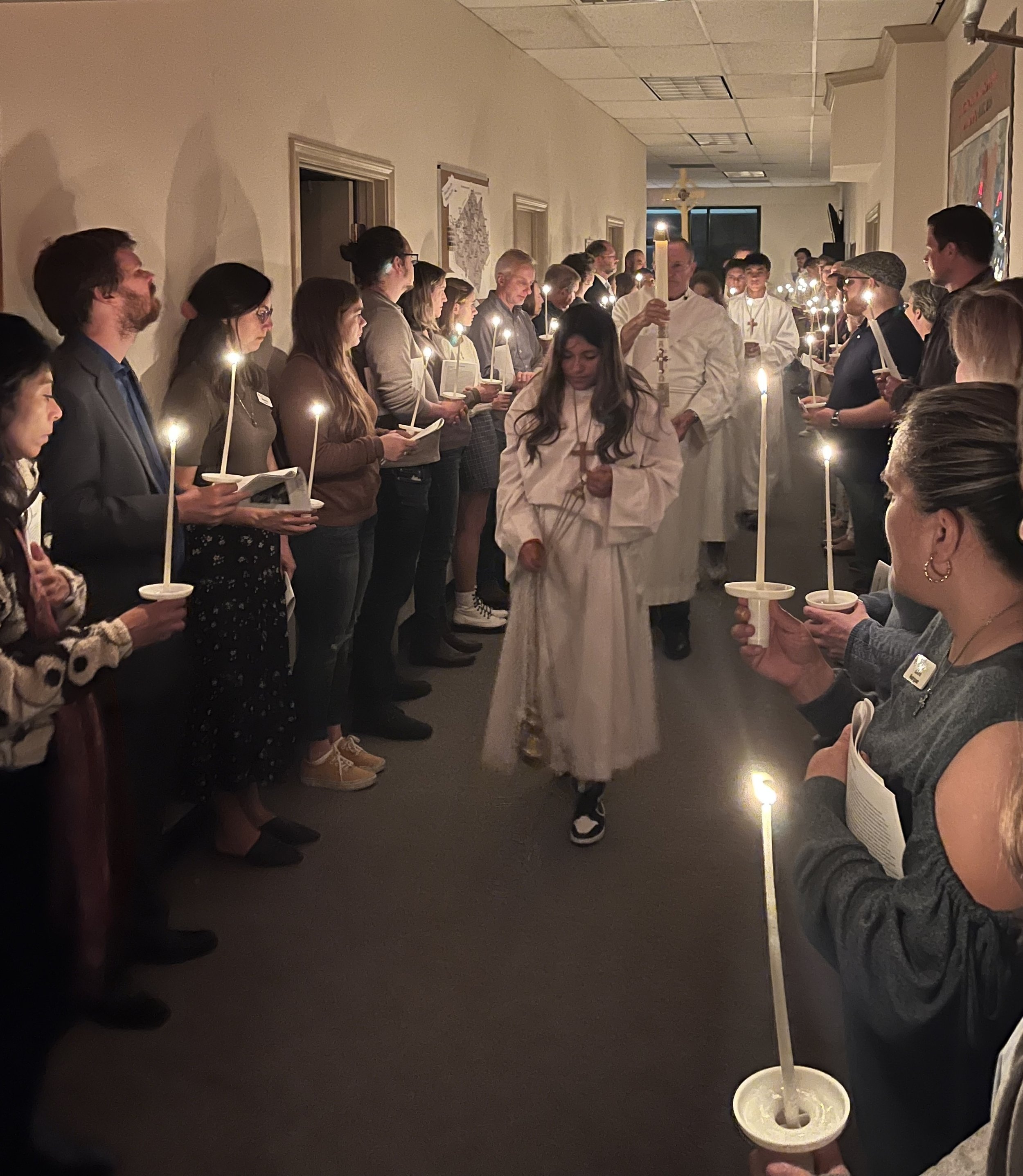 People participating in a candlelight procession inside a building, with some wearing white robes and others holding lit candles in paper holders, and a person with a cross walking at the front.