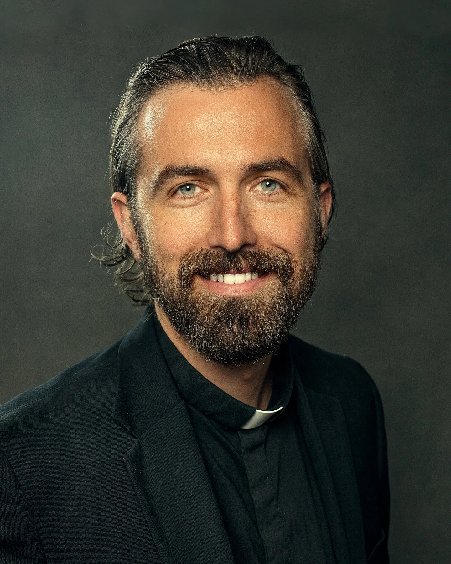 A portrait of Fr. Kurt Hein with a beard and mustache, wearing a black clerical shirt with a white collar, against a dark background.