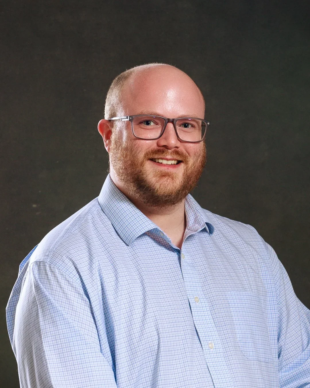 A man with glasses and a beard in a light blue checked shirt smiling for a portrait against a dark background.