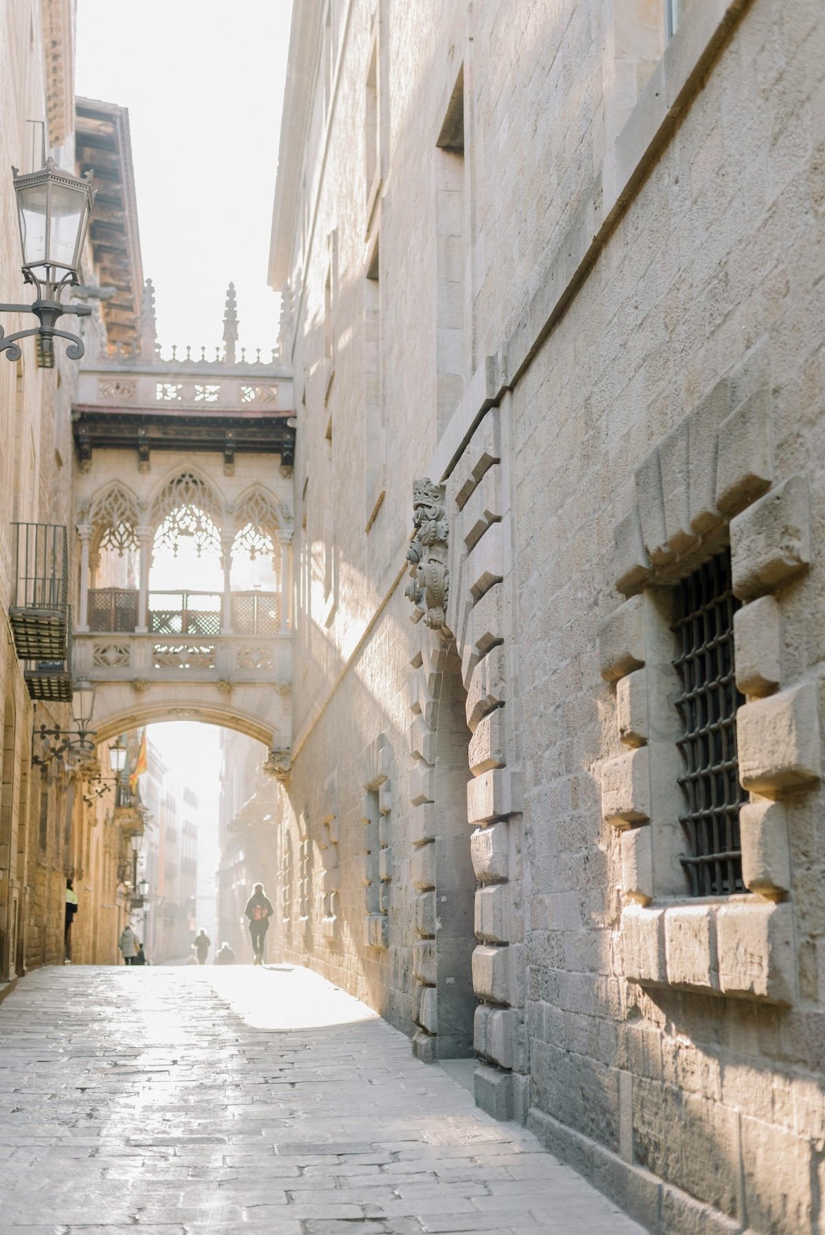 A narrow cobblestone street with historic stone buildings, decorative balconies, and an ornate bridge connecting two buildings, with a few pedestrians walking in the distance.