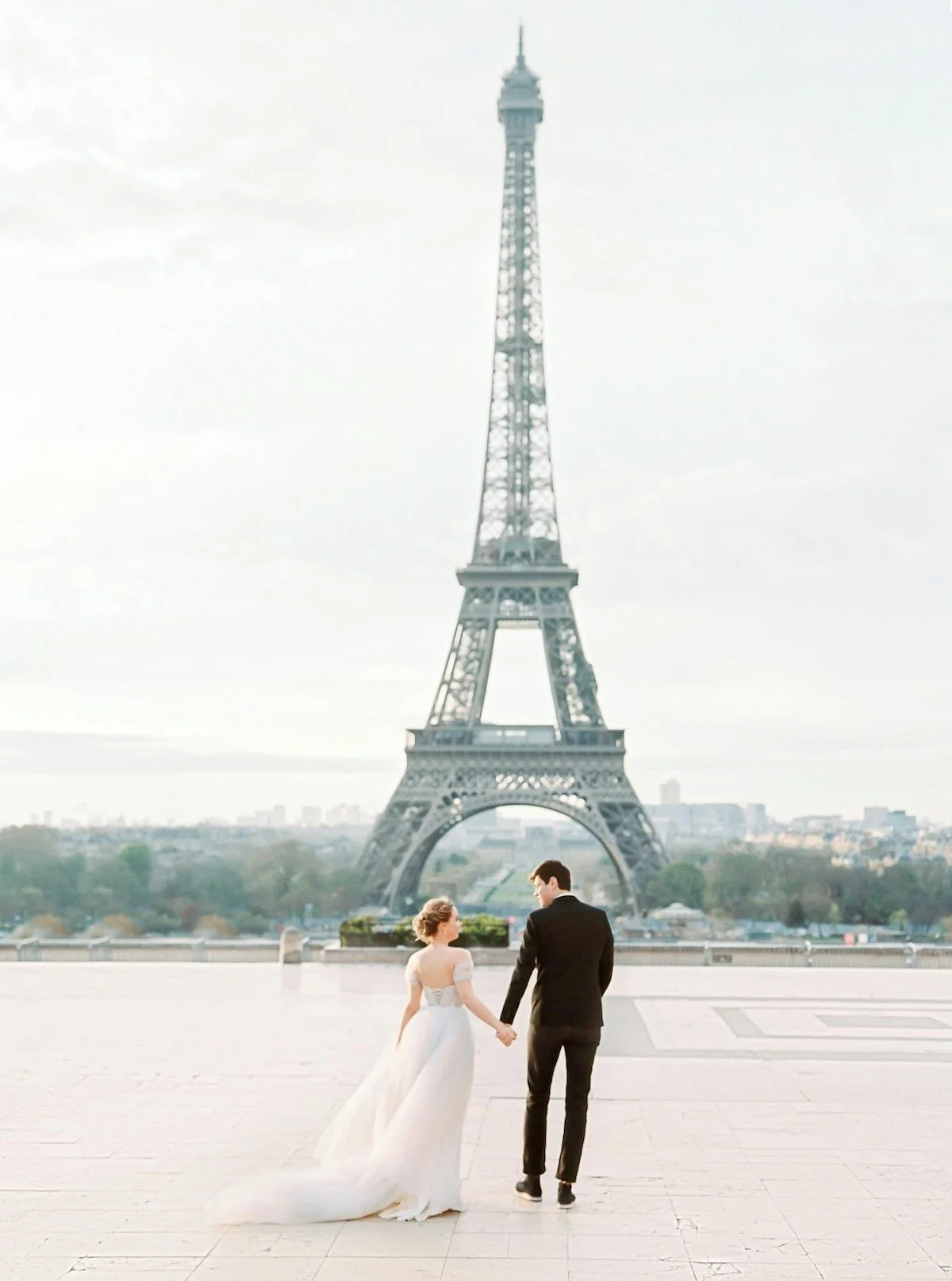 A bride in a white wedding dress and a groom in a black suit holding hands, walking together in front of the Eiffel Tower in Paris.