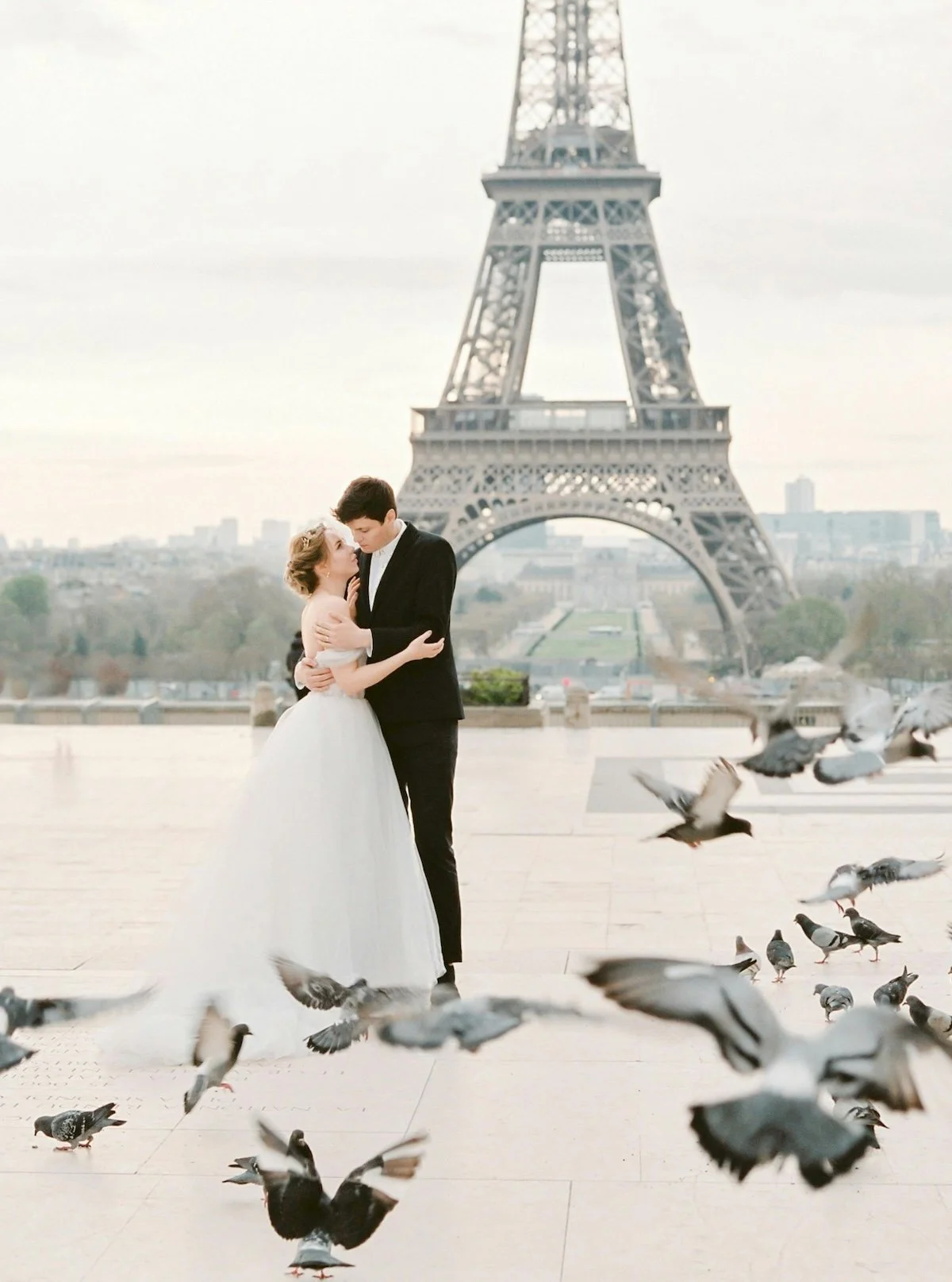 A couple in wedding attire, with the bride in a white gown and the groom in a black suit, embracing in front of the Eiffel Tower during daytime, with pigeons flying around.