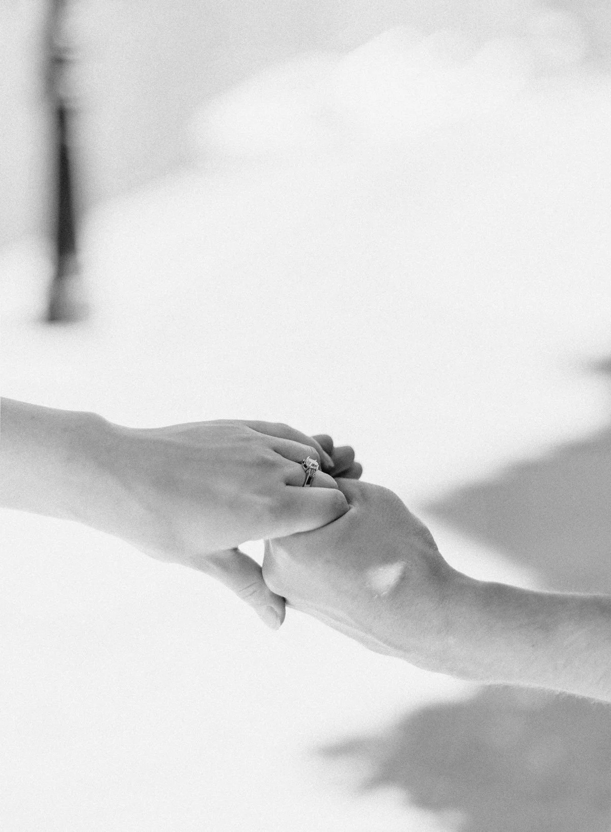 Close-up of two hands clasped, one with a wedding ring, on a neutral background.