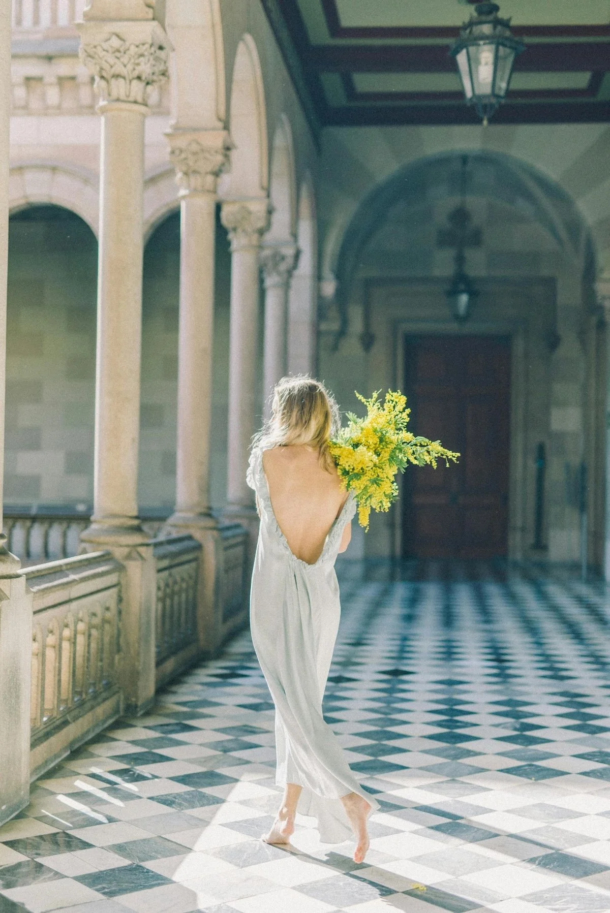 Woman in a backless satin dress holding a bouquet of yellow flowers, walking across a black-and-white checkered floor in a historic building with stone columns and arches.