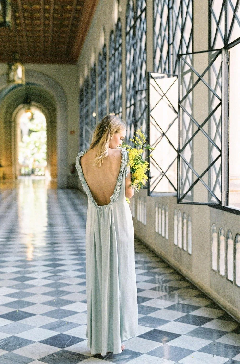 A woman in a white dress with an open back, holding a bouquet of yellow flowers, standing in a bright corridor with checkered floor and large windows.