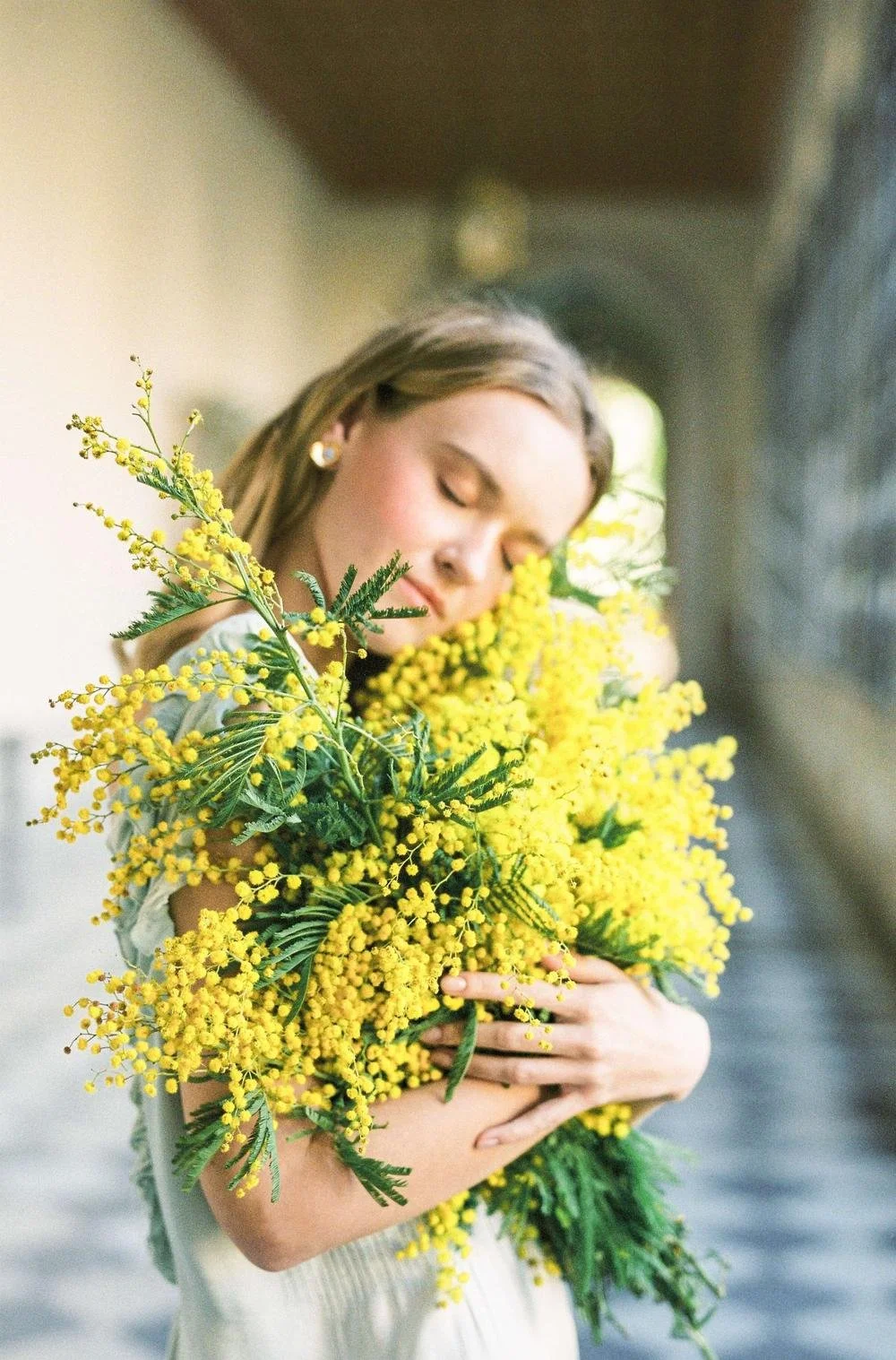 A woman with light brown hair and earrings is holding a large bouquet of yellow flowers with green leaves, her eyes closed and face relaxed.