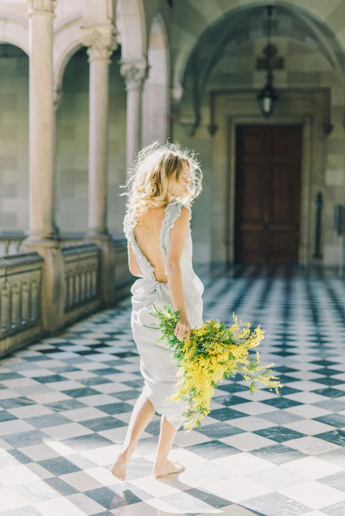 A woman with curly blonde hair, dressed in a white gown with open back details, holding a large yellow flower bouquet, walking barefoot in a grand, historic hall with checkered black and white floors and ornate stone columns.