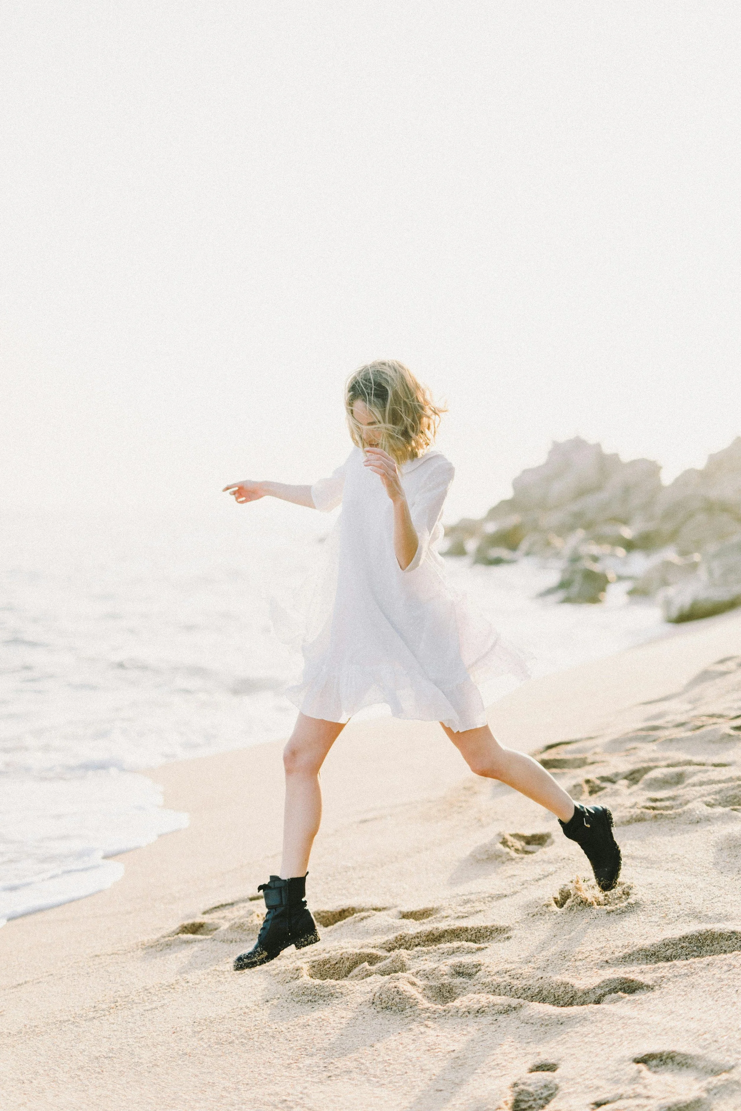 A young girl running along the sandy beach near the ocean, with rocks in the background and her hair flowing in the wind, wearing a white dress and black boots.
