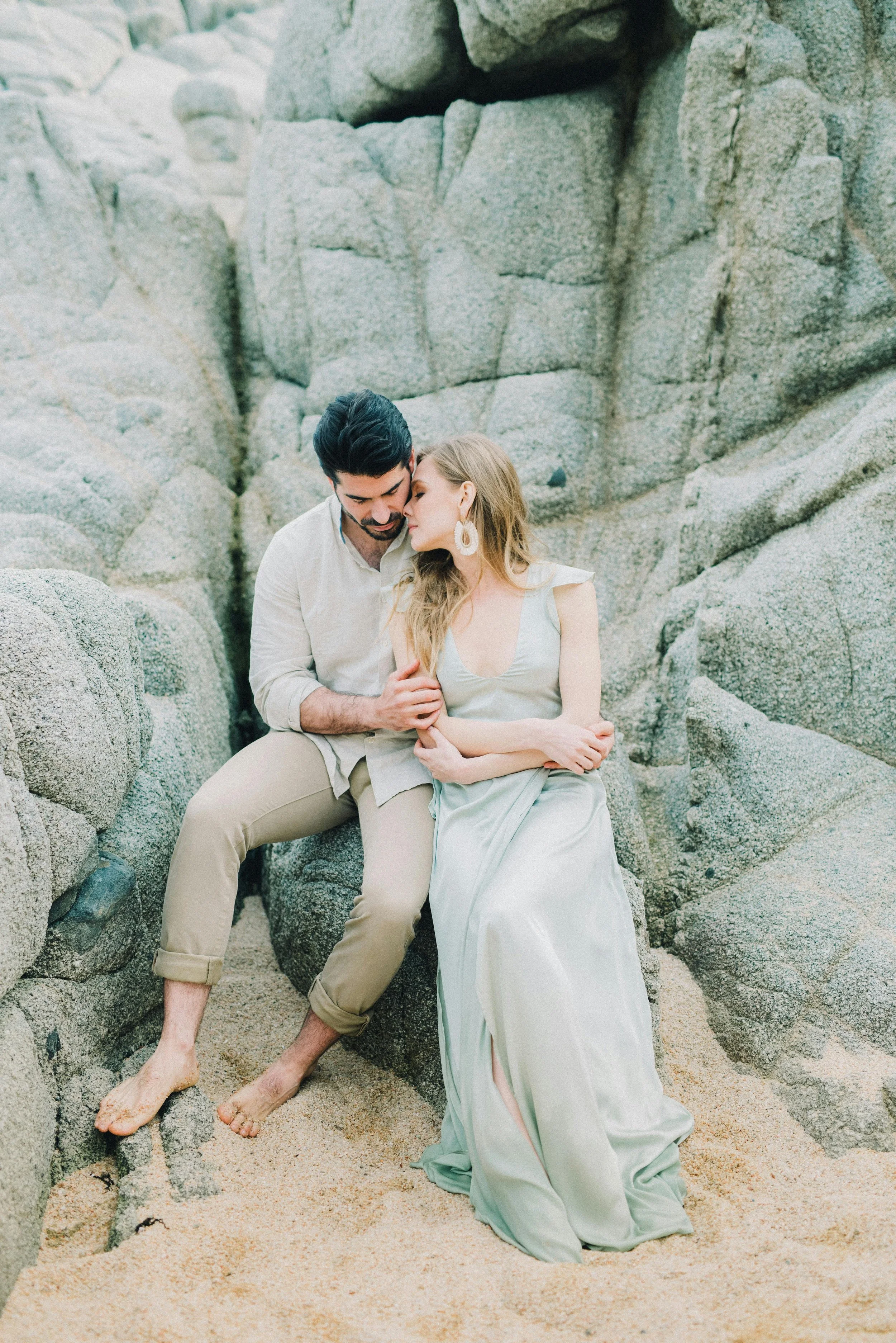 A couple sitting close together on rocks at the beach, with the man touching the woman's arm. The woman has long blonde hair and is wearing a light-colored dress and large earrings. The man has dark hair and a beard, wearing a light shirt and rolled-up pants, both barefoot. The scene appears intimate and serene.
