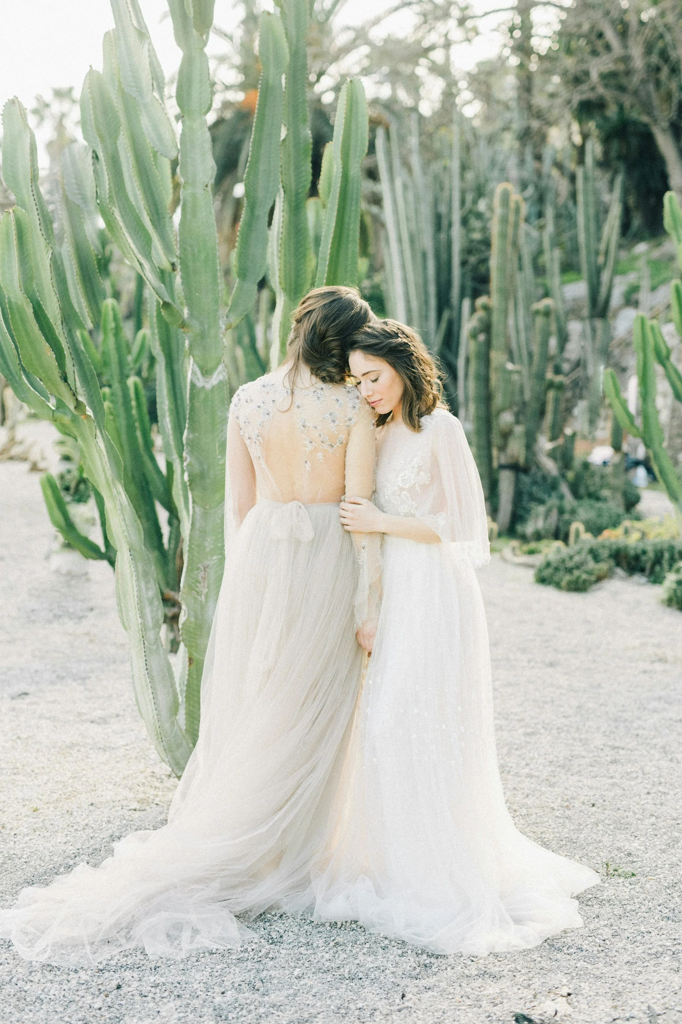 Two women in flowing wedding dresses sharing an intimate moment in front of large cacti in a desert garden.